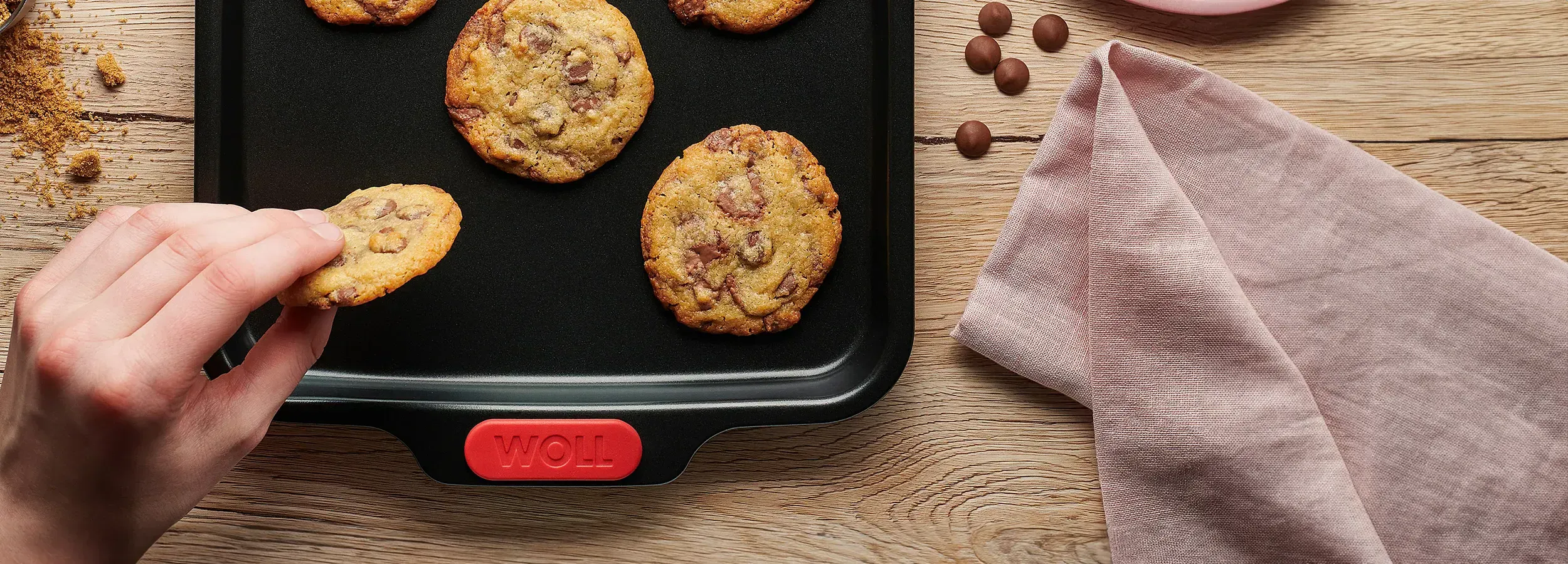 Auf einem Backblech der Serie Let`s Bake! liegen frischgebackende Cookies, als Deko liegen Schokodrops, eine Servierte auf dem Tisch.