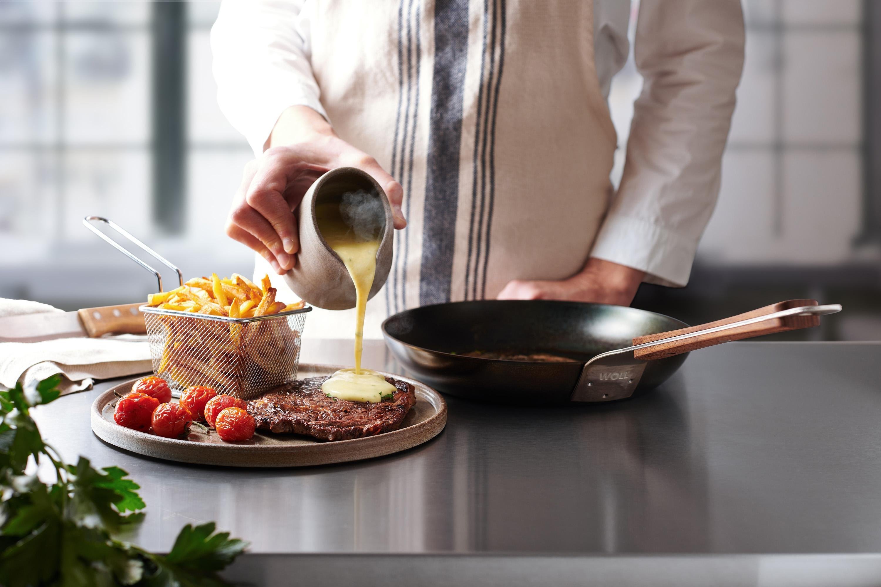 Carbon Steel Flachpfanne Auf einem Teller liegt ein angebratenes Steak, das mit Soße übergossen wird. Daneben steht eine runde Carbon Steel Pfanne.