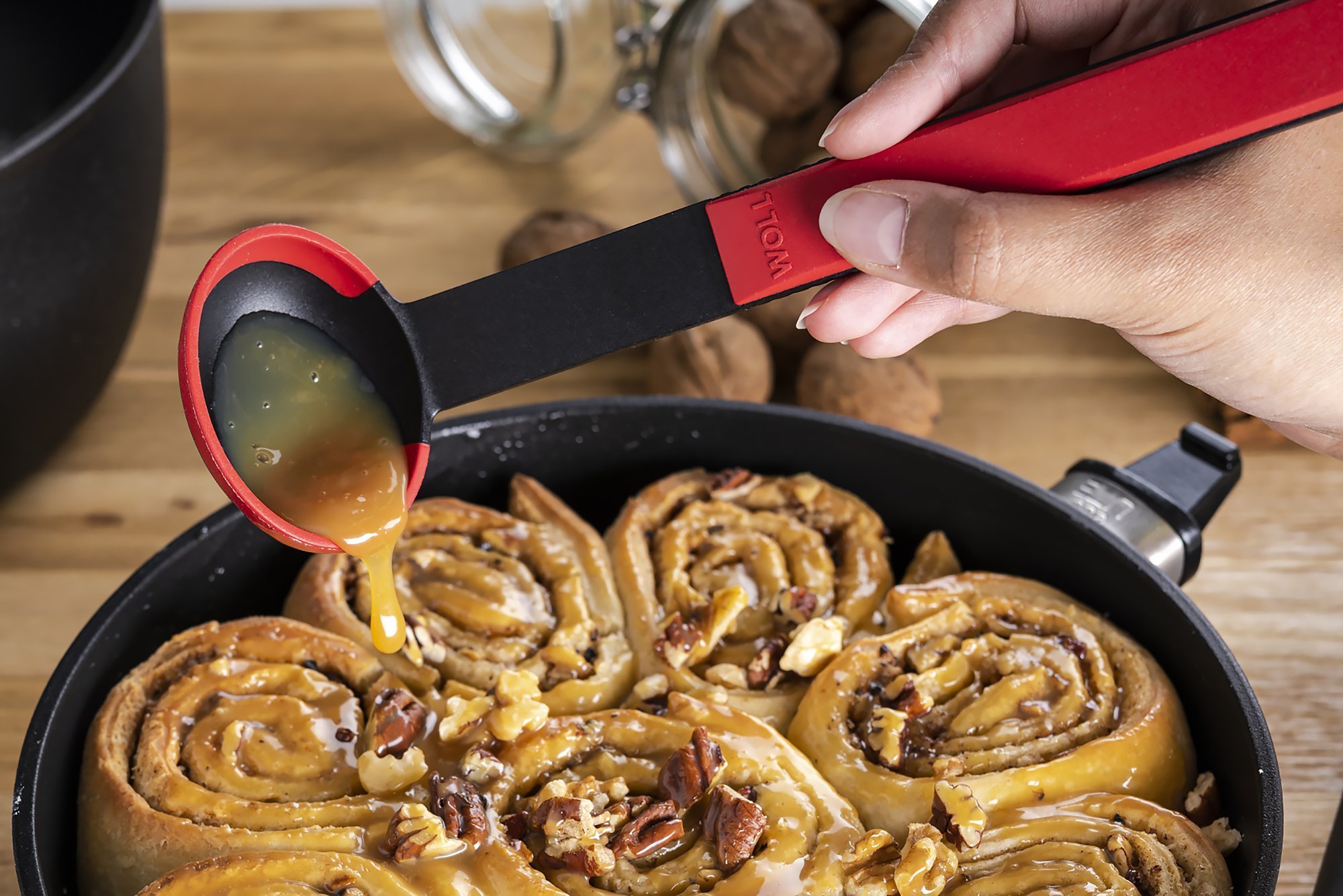 In einer runden Eco-Lite-QXR-Pfanne befinden sich  Zimtschnecken, diese werden mit einem roten Cock it Saucenlöffel mit Karamellsoße übergossen. 