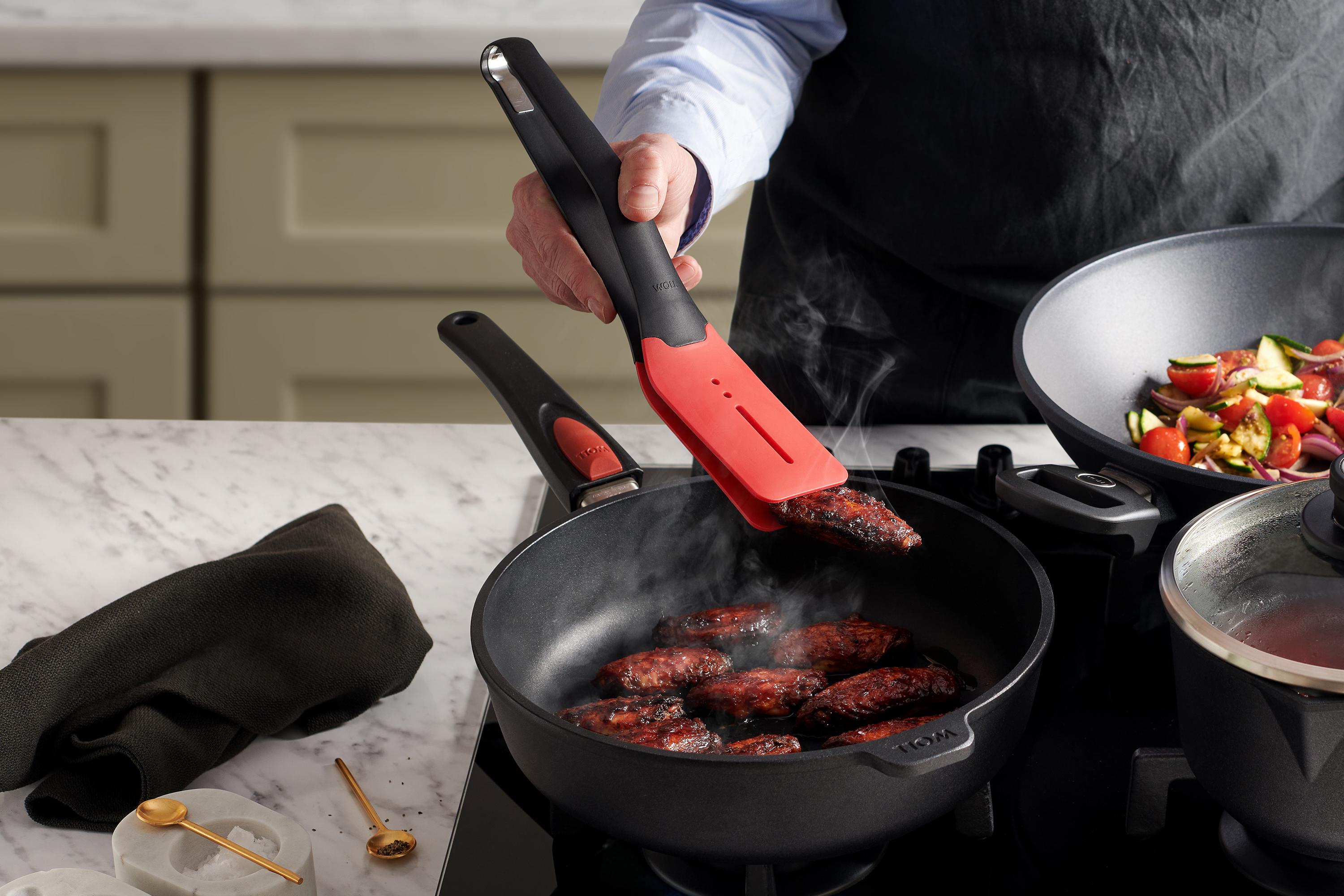 Titan Best Guss-Hochrandpfanne In einer runden Titan Best Pfanne werden Fleischstücke angebraten und mit einer Cock it Küchenzange gewendet.