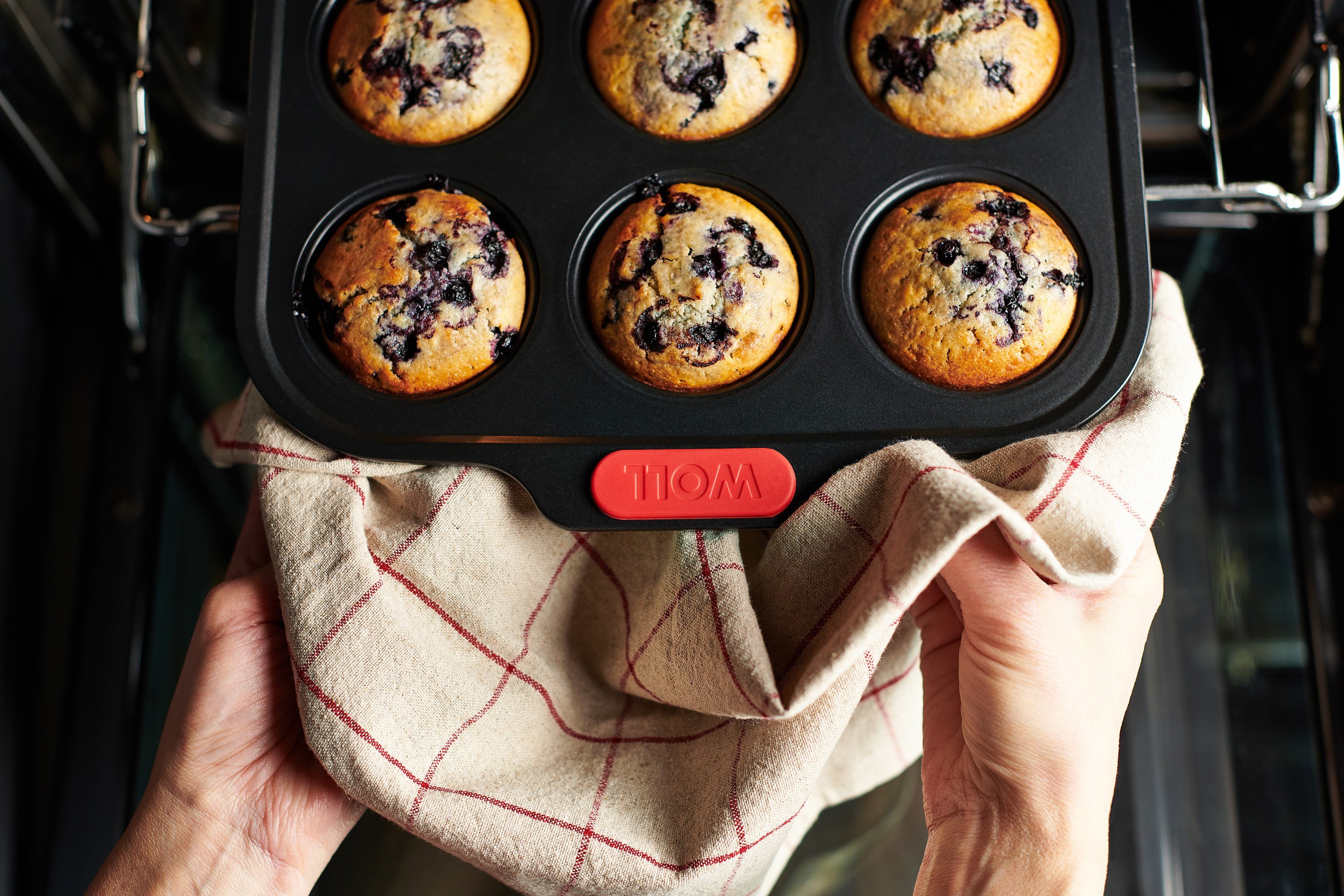 Eine heiße rechteckige Muffinform der Serie Let's Bake! enthält frisch gebackene Blaubeermuffins. Sie wird mit einem Küchentuch aus dem Backofen genommen.