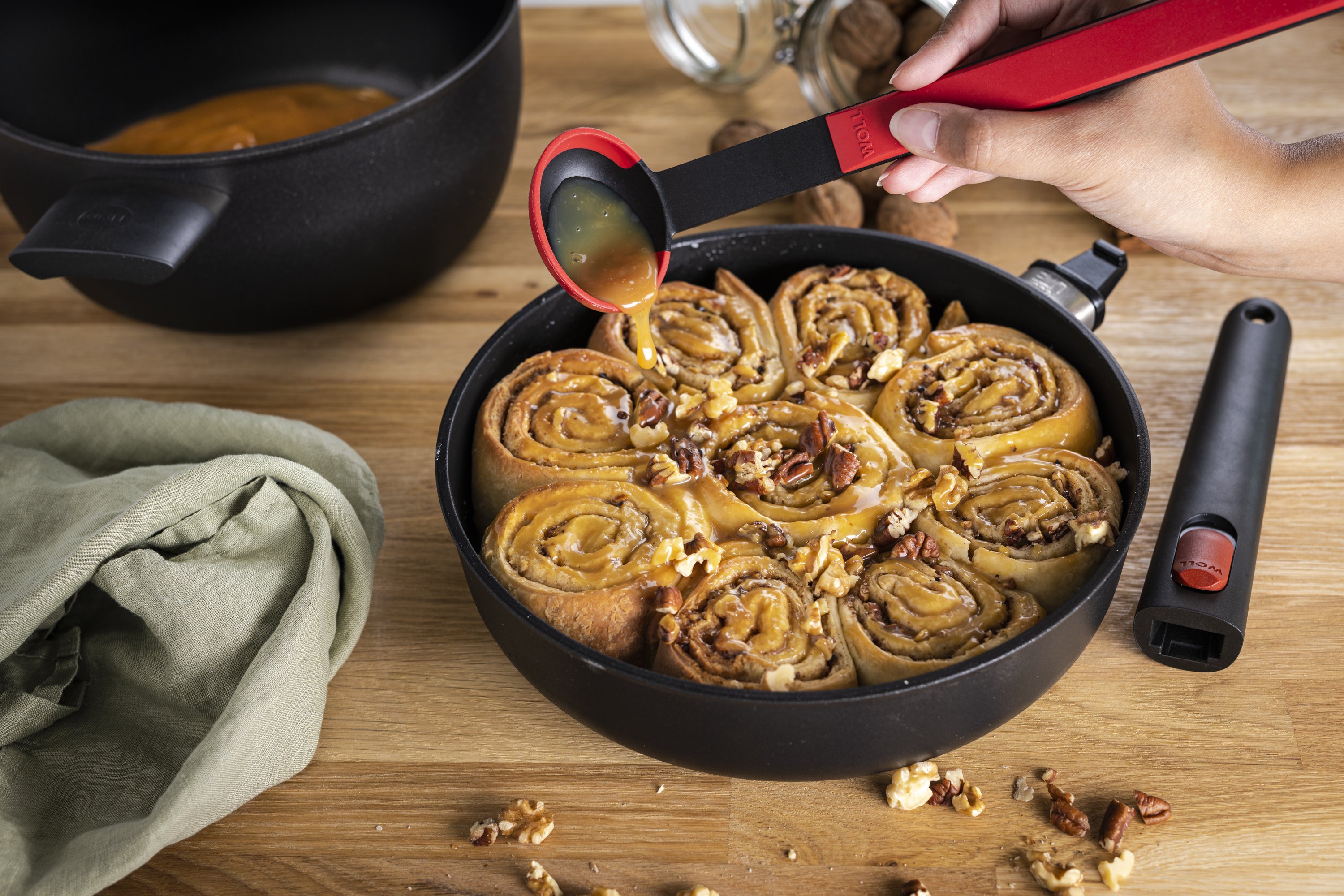 1724ELI_124ELI_Zimtschnecken_3000x2000px In einer runden Eco-Lite-Pfanne ohne Stiel befinden sich frisch gebackene Zimtschnecken. Die Zimtschnecken sind mit Walnussplittern garniert und werden mit einem Saucenlöffel mit Karamellsoße übergossen. Im Hintergrund steht ein runder Topf, in dem die Ka