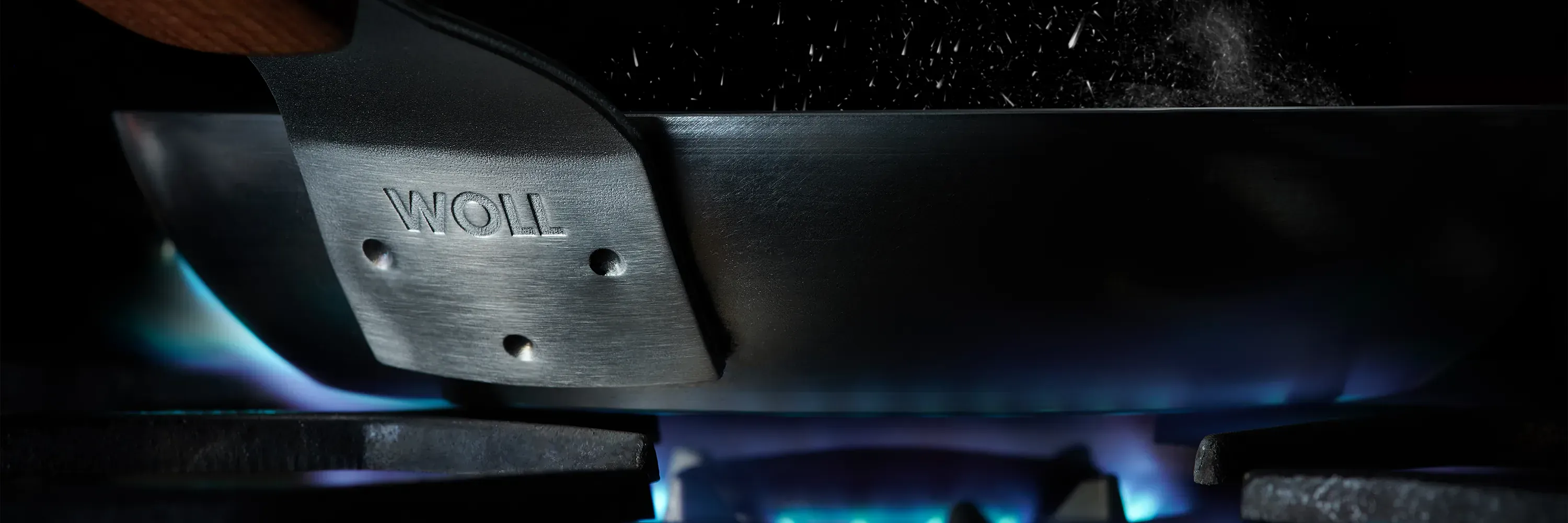 In einer Nahaufnahme sind die Befestigungspunkte des Griffs an der Carbon-Steel-Pfanne zu erkennen. Die Pfanne steht auf einem Gasherd, darunter brennen blaue Flammen.