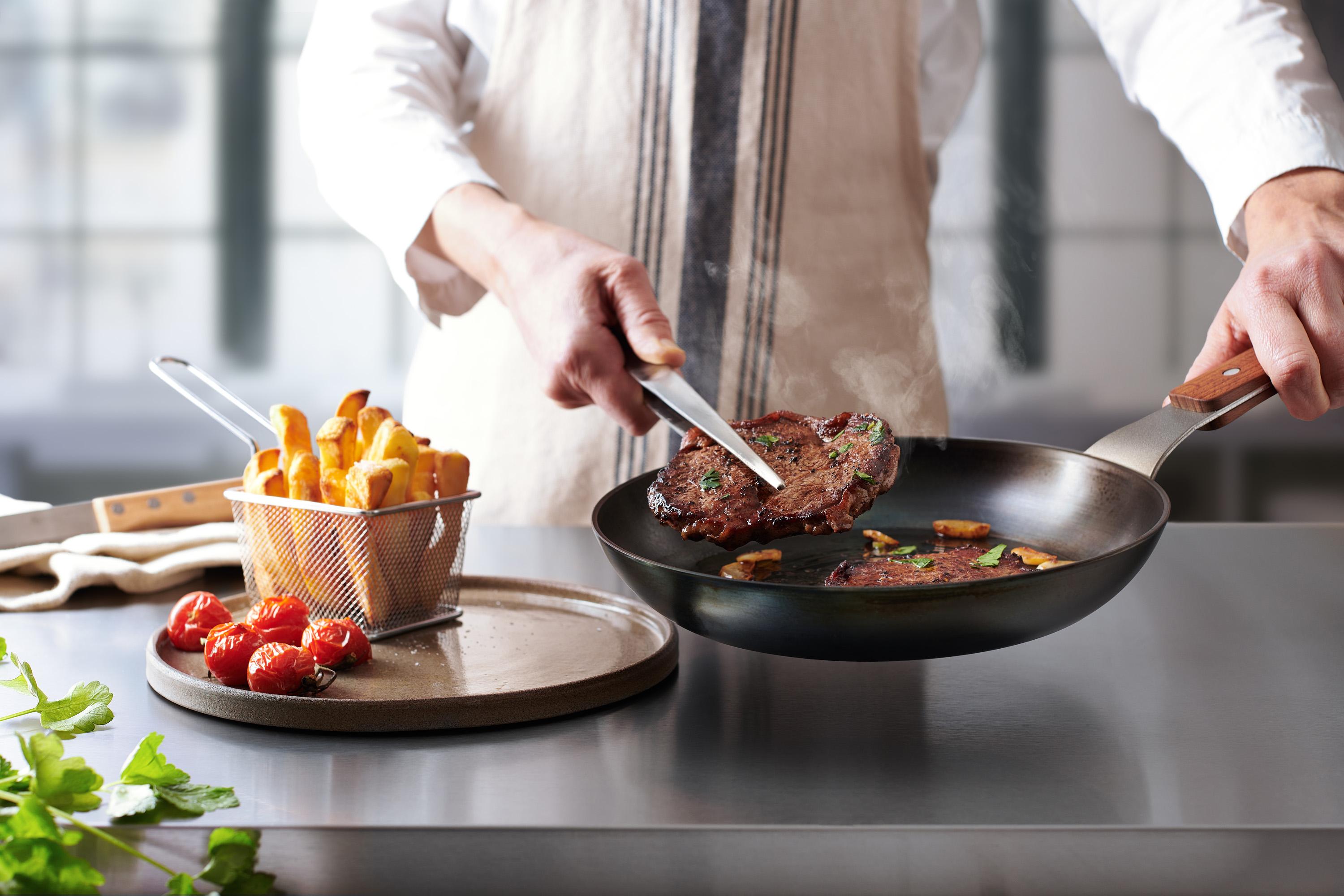 Carbon Steel Flachpfanne In einer runden Carbon Steel Pfanne befinden sich angebratene Steaks. Ein Steak wird auf einem Teller platziert.
