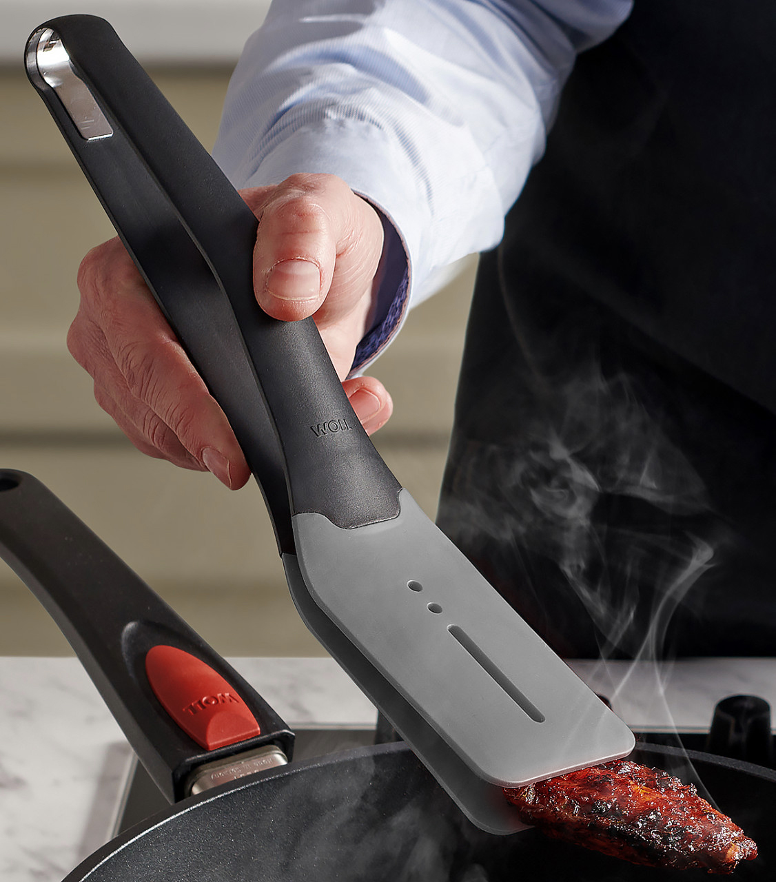 Cook it Küchenzange Ein dampfendes, angebratenes Stück Fleisch wird mithilfe der grauen Cock it Küchenzange in der heißen Pfanne gewendet.