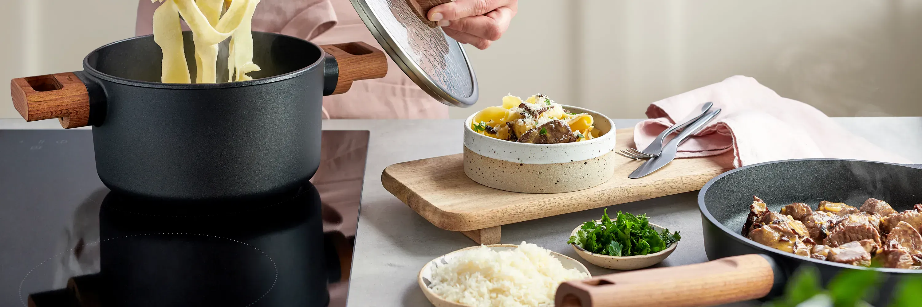 Im Topf der Serie Eco Logic QXR wurden frische Nudeln gekocht, die nun entnommen werden. Dazu wurde der Deckel abgenommen. Auf der Arbeitsplatte steht ein Holzbrett mit einem angerichteten Pastateller. Daneben liegen Besteck und eine Serviette. Um den Tel