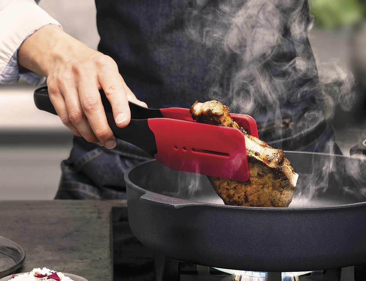 Ein dampfendes, angebratenes Stück Fleisch wird mithilfe der roten Cock it Küchenzange in der heißen Pfanne gewendet.