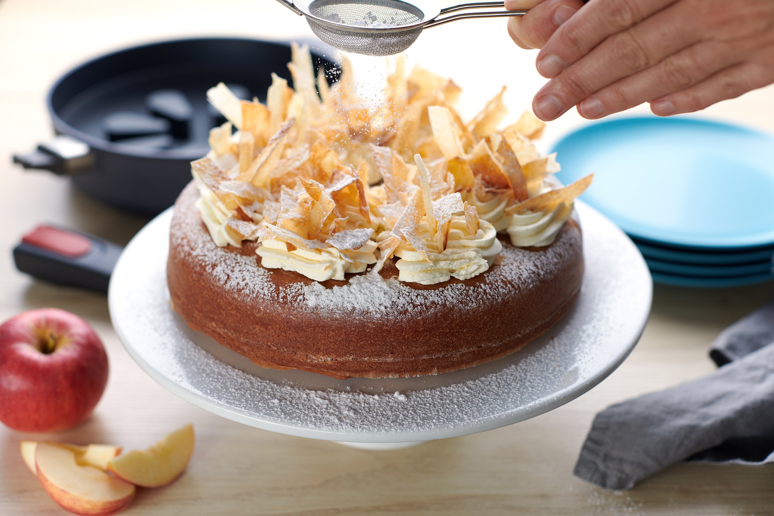 Auf einer Kuchenplatte steht ein frisch gebackener Apfelkuchen, der mit Puderzucker bestäubt wird. Im Hintergrund ist eine runde Diamond-Lite-Pfanne mit abgenommenem Stiel zu sehen. In dieser befindet sich ein Silikon-Backeinsatz. Drumherum stehen noch Te