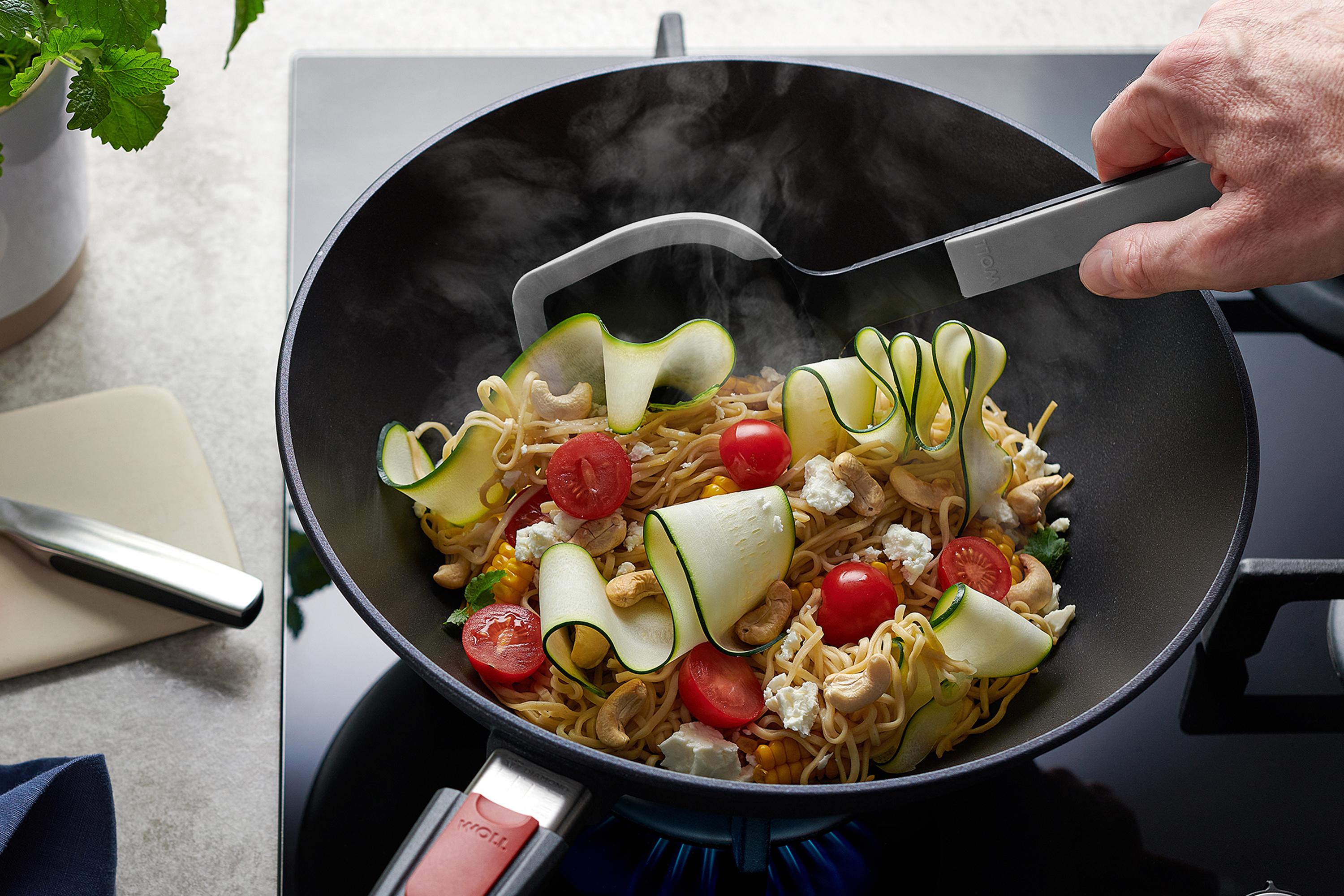 Cook it Wok-Wender In einer runden Diamond Lite Wok- und Rührpfanne wird ein Nudelgericht zubereitet, das mit einer grauen Cock it Pfannenschaufel umgerührt wird.
