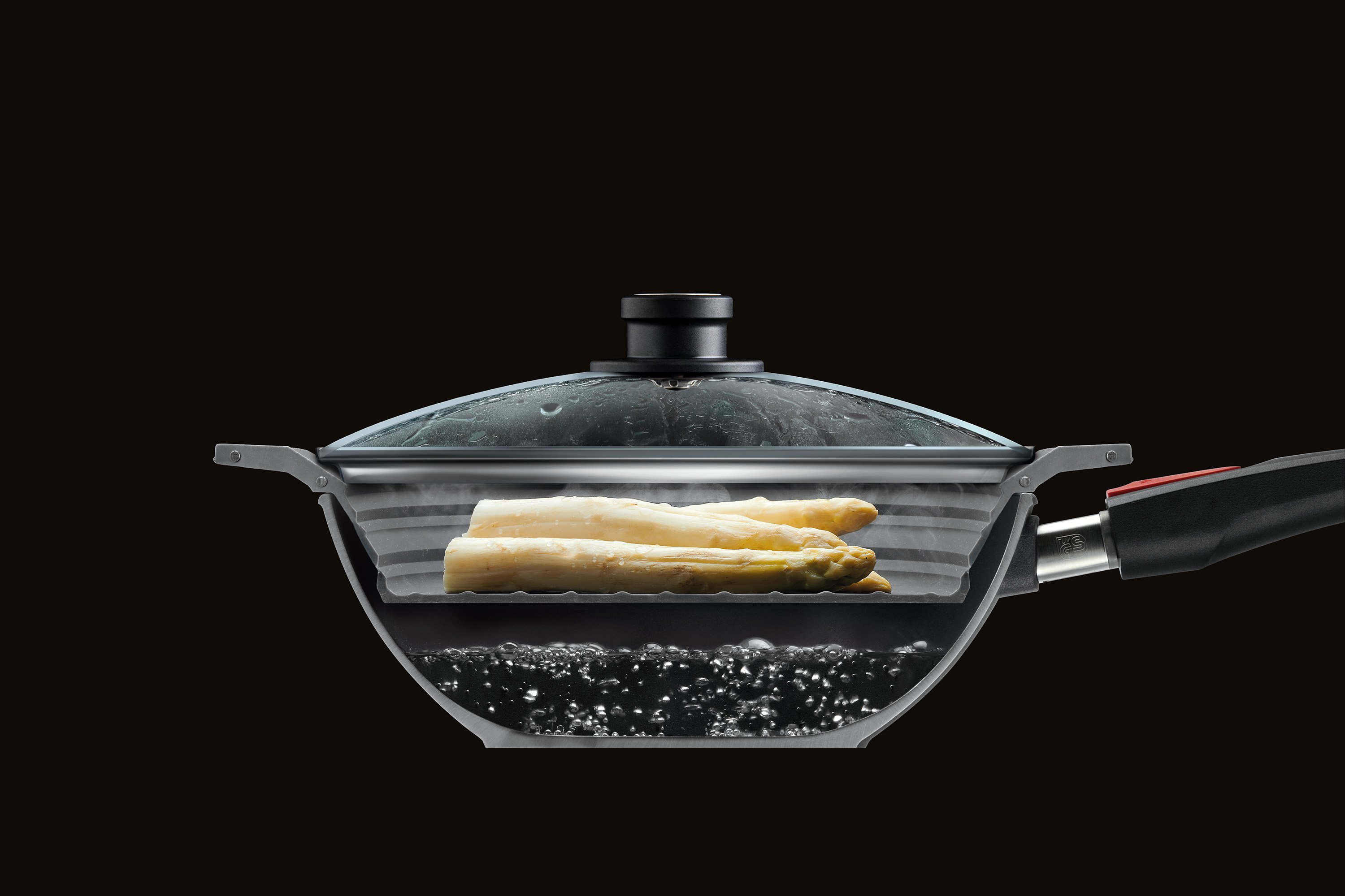 Im Querschnitt ist eine halbierte Wok Pfanne mit Glasdeckel zu sehen, in der Spargel mit dem Steam it multifunktionalen Silikoneinsatz gedämpft wird. 