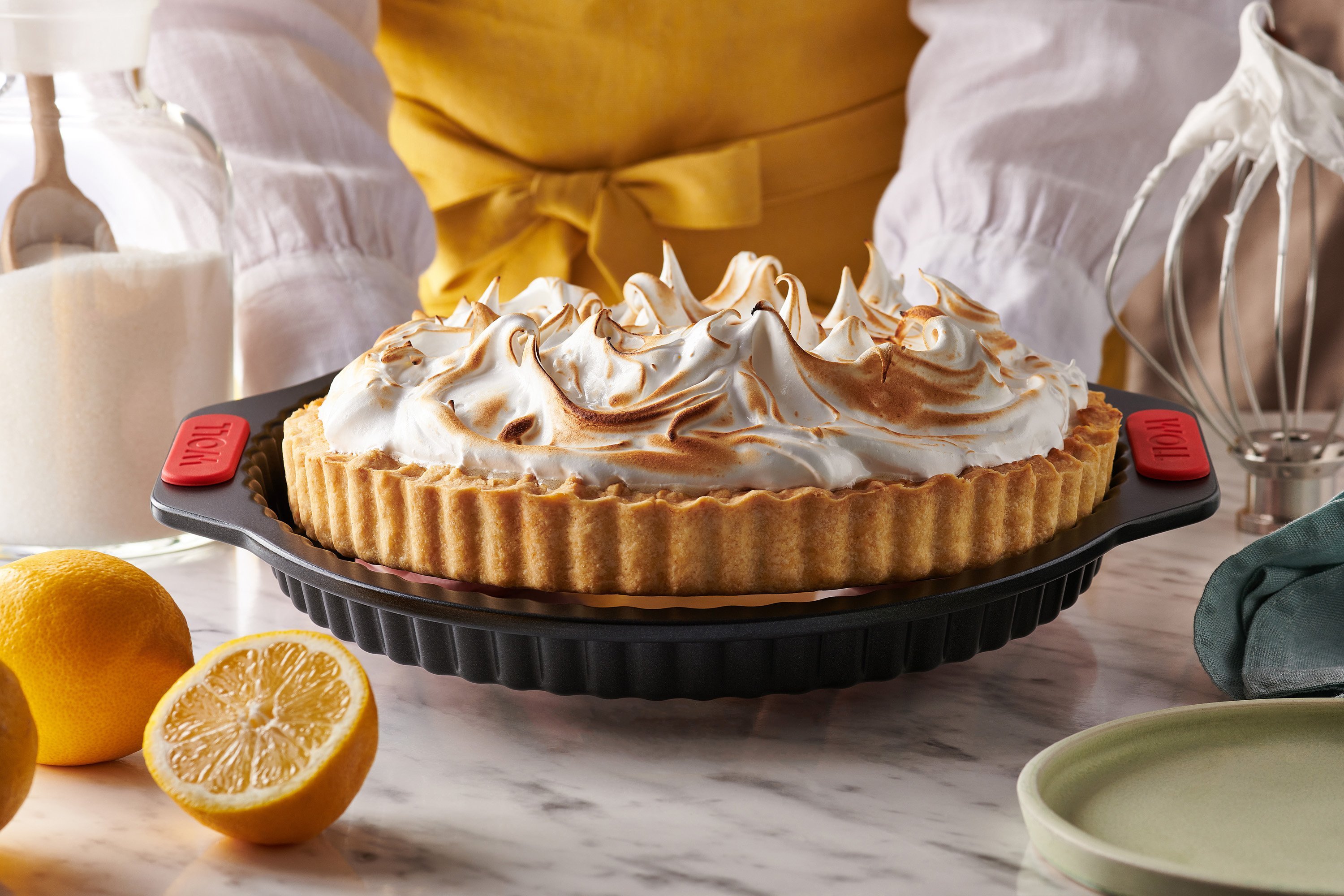 Auf einem Tisch steht eine Quiche- und Tarteform mit Hebeboden aus der Serie Let's Bake! und enthält eine frisch gebackene Zitronen-Baiser-Torte. Daneben liegen aufgeschnittene Zitronen.