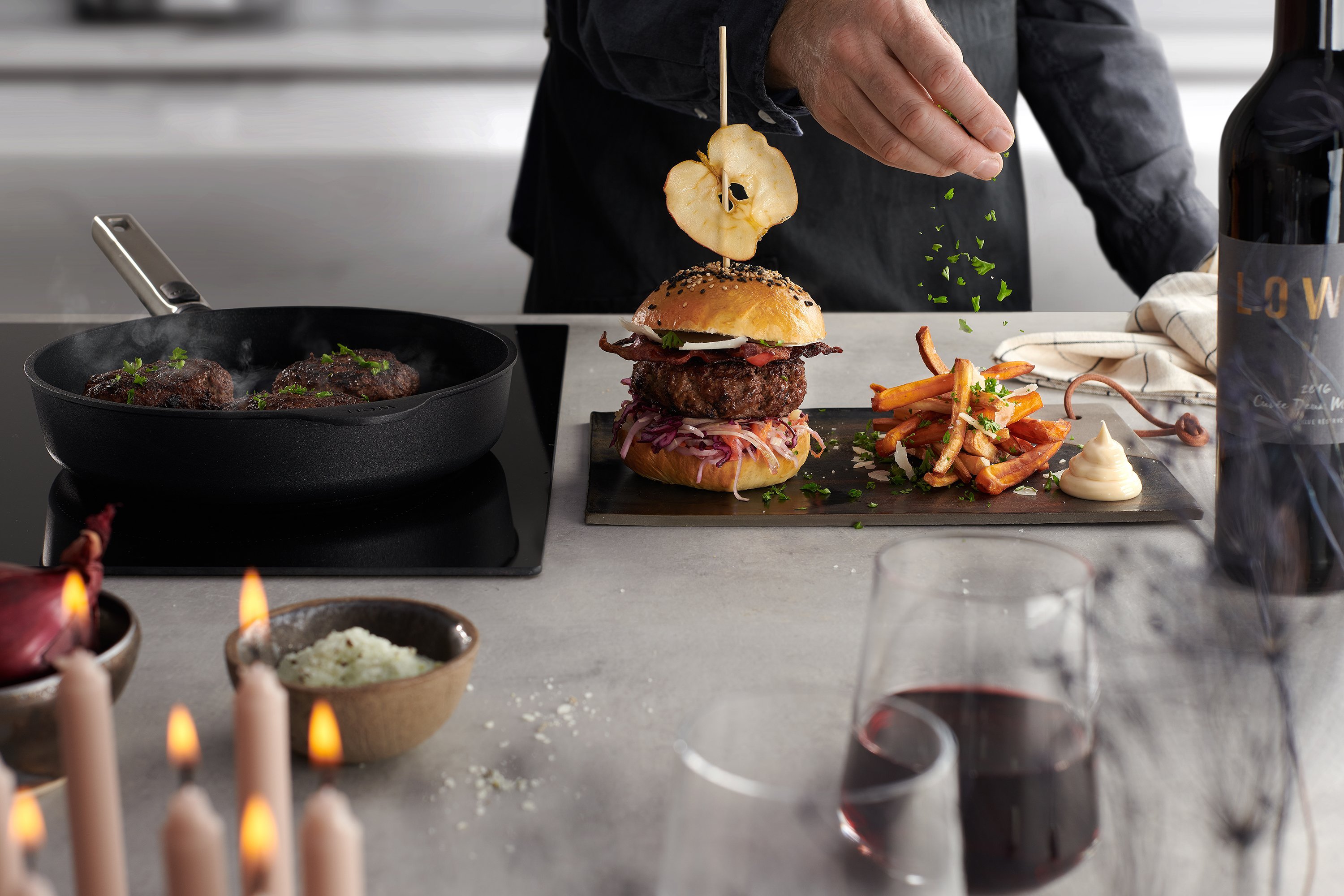 Die Burgerpattys werden in einer heißen, runden Logic-Pfanne mit abnehmbarem Edelstahlstiel angebraten. Ein Burger in einem hausgemachten Brötchen mit eingelegtem Krautsalat und Süßkartoffel-Pastinaken-Chips wurde auf einer Platte angerichtet.