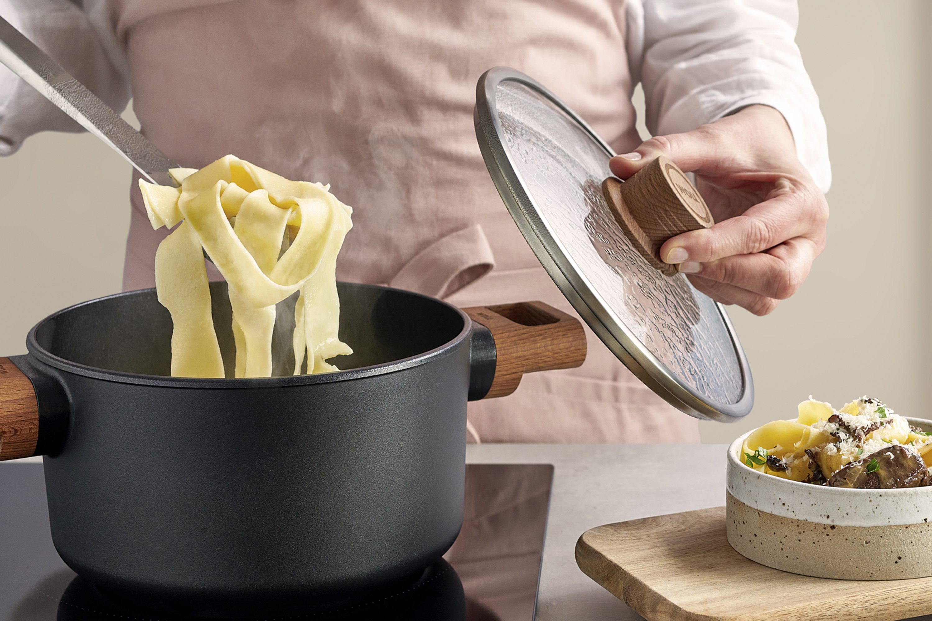 In einem runden Eco Logic QXR Topf werden gekochten Nudeln entnommen, während der Glasdeckel mit einer Hand festgehalten wird. 