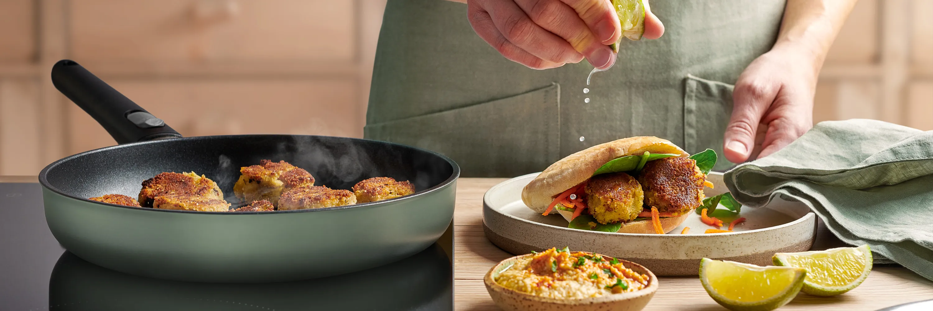 In einer runden, heißen Ecolour Pfanne werden frische Falafel angebraten. Auf dem Teller daneben liegt ein fertiges Fladenbrot gefüllt mit Falafel und eine Schüssel Hummus steht vor dem Teller.
