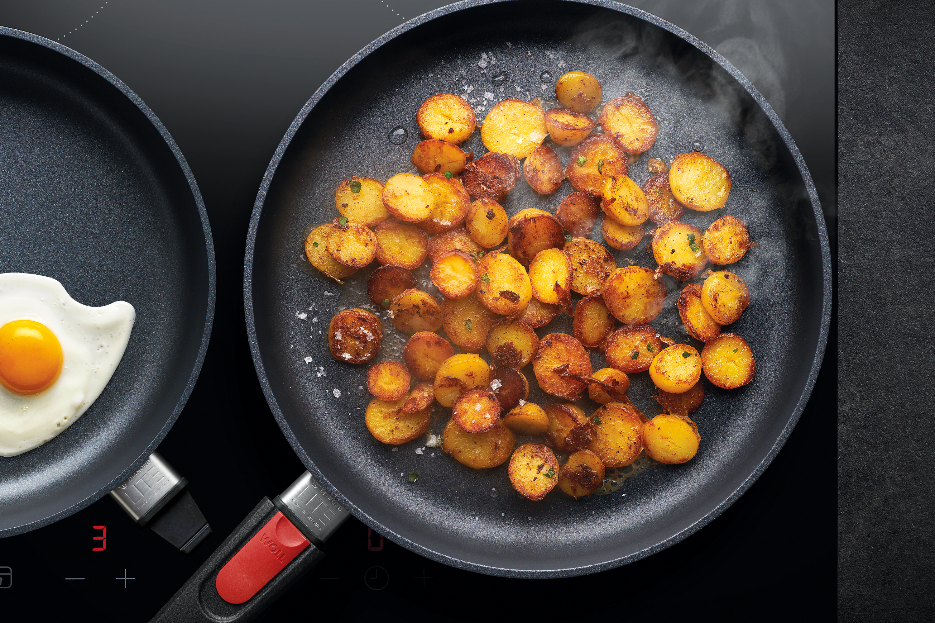 Diamond Lite Gusspfanne In einer runden Diamond Lite Pfanne werden Bratkartoffeln angebraten, während in einer weiteren Pfanne Spiegeleier zubereitet werden.