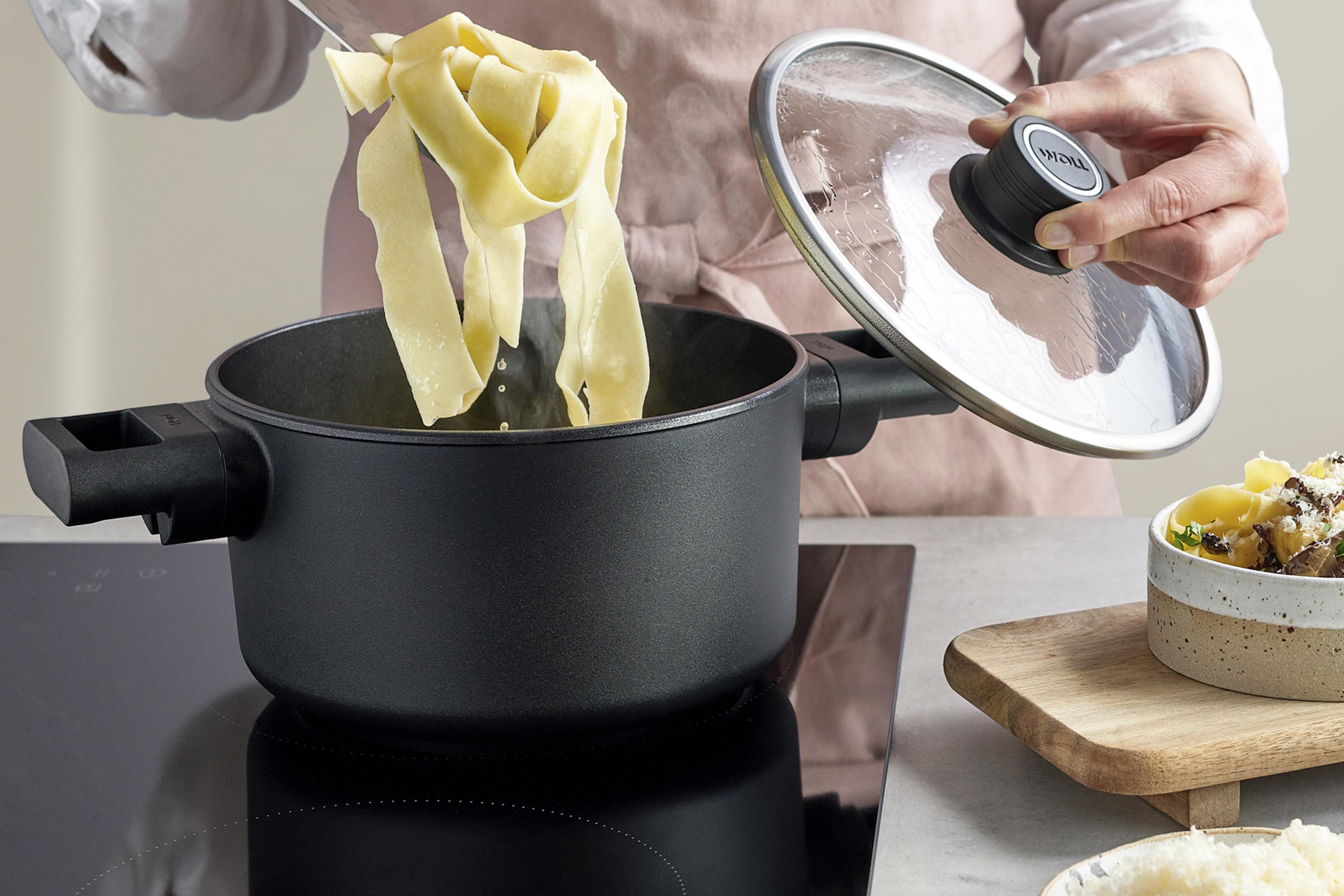 In einem runden Eco Logic QXR Topf werden gekochten Nudeln entnommen, während der Glasdeckel mit einer Hand festgehalten wird. 