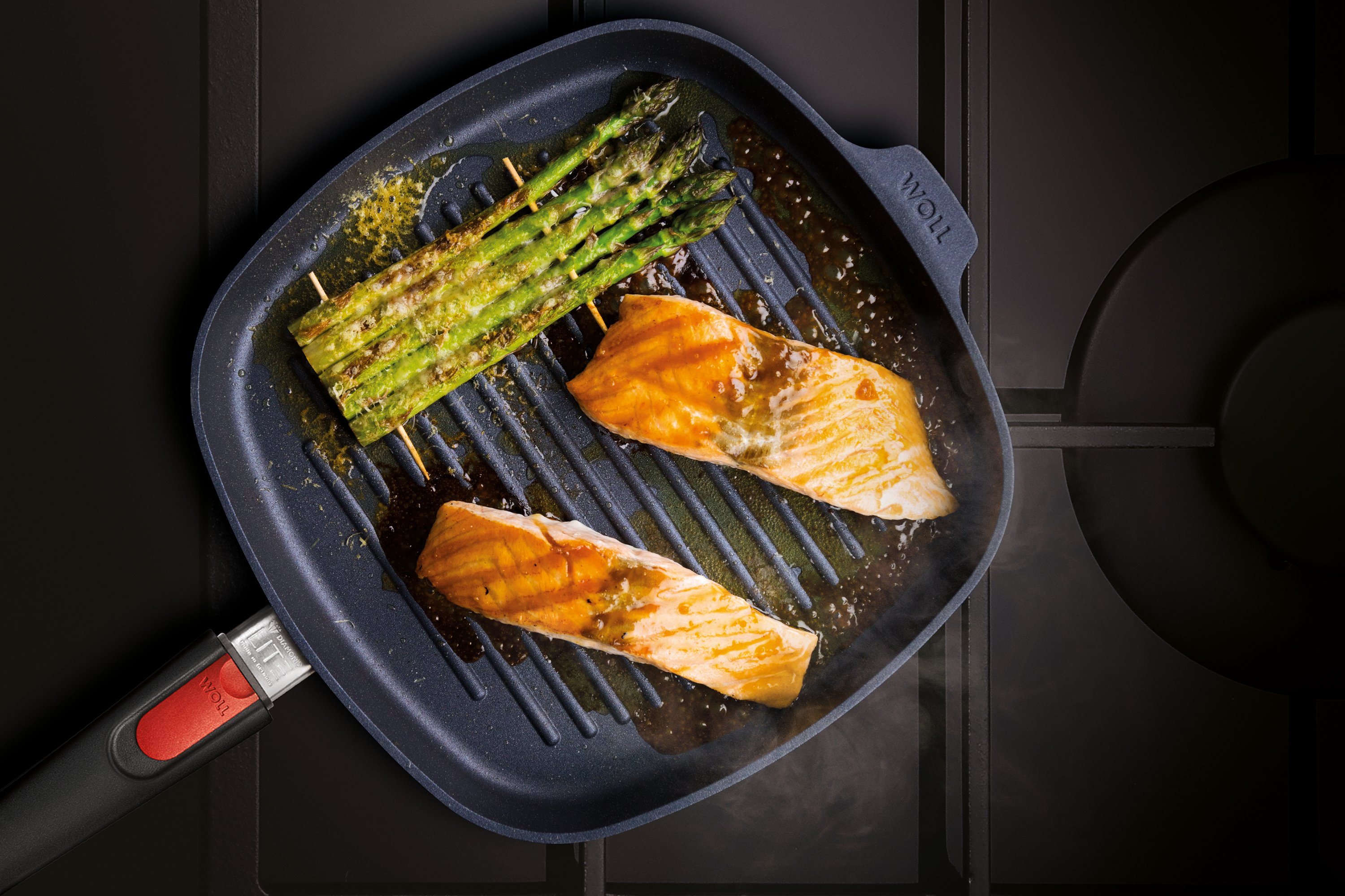 Auf dem Herd steht eine viereckige, heiße Diamond-Lite-Grillpfanne mit abnehmbarem Stiel. Darin befindet sich frisch zubereiteter, dampfender Honig-Senf-Lachs mit grünem Spargel.