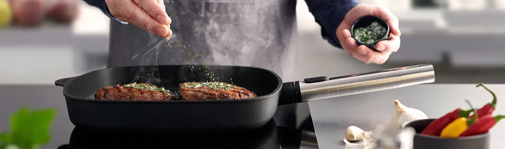 Auf dem Herd steht die rechteckige, heiße Logic-Grillpfanne mit abnehmbarem Edelstahlstiel, in der saftig angebratenes Fleisch dampft. Es ist mit Kräutersalz bestreut. Daneben liegen frische Knoblauchzehen auf der Arbeitsplatte und in einem Behälter stehe