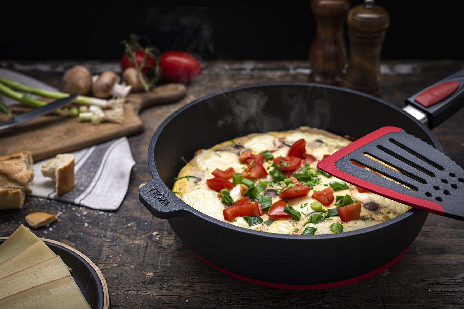 Auf dem Tisch steht eine runde, heiße Titan-Best-Pfanne mit abnehmbarem Stiel. In dieser befindet sich ein frisches Raclette-Omelett. Auf dem Pfannenrand liegt ein Jumbo-Pfannenwender aus der Serie Cook it.
