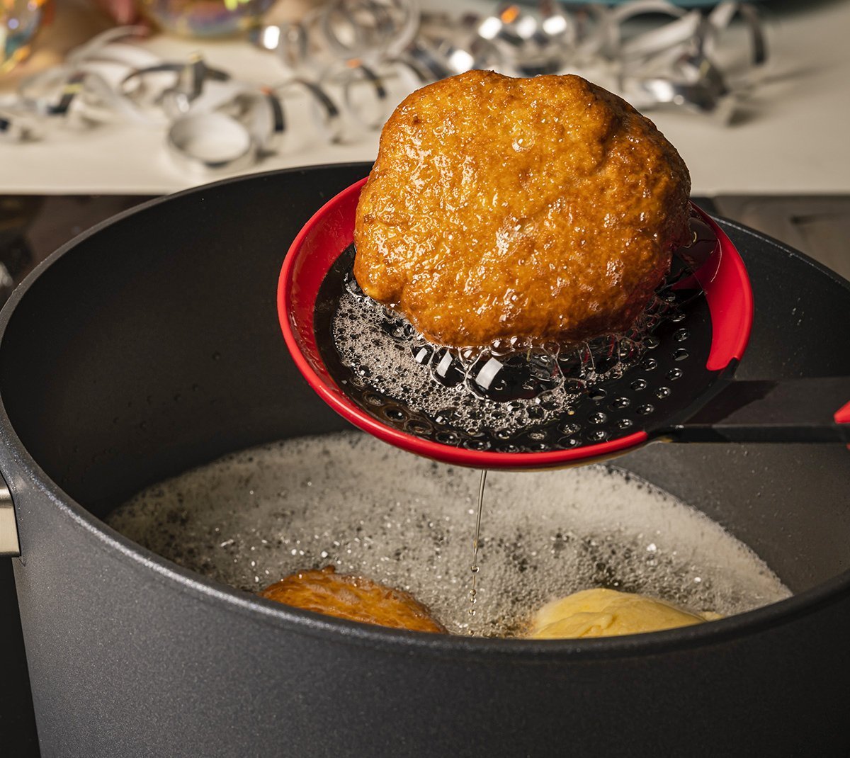Mit einer roten Cock it Schöpfkelle wird ein ausbackener Fastnachtkrapfen aus dem Topf entnommen und über dem Topf abgetropft.