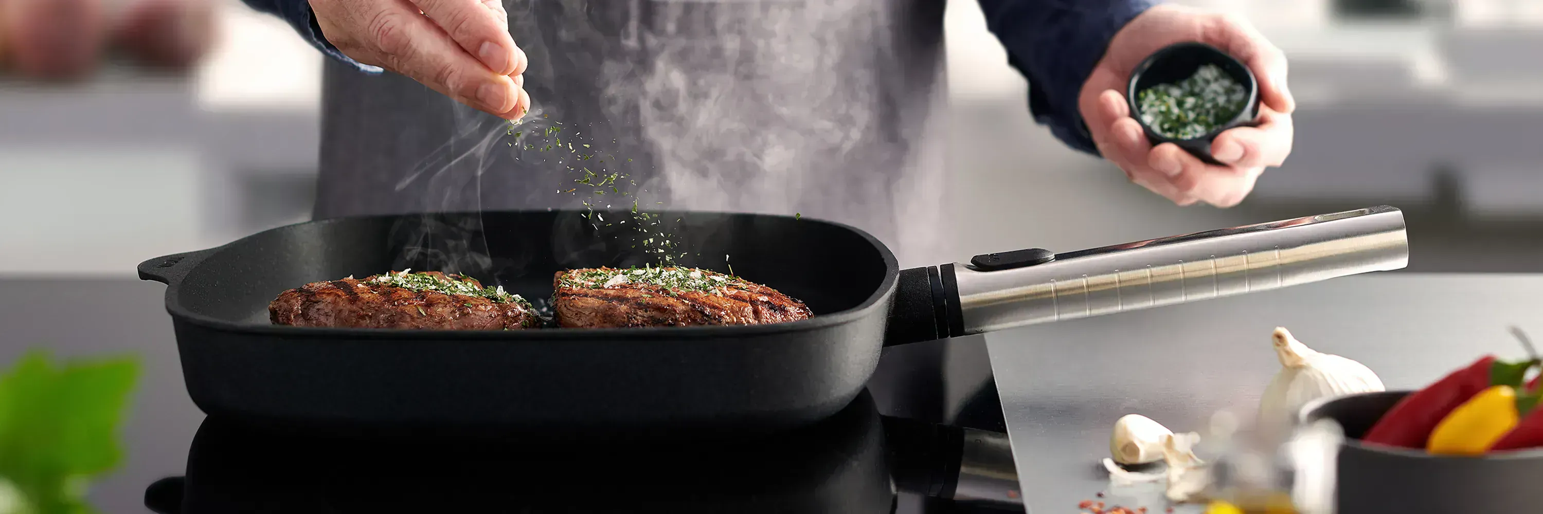 In einer viereckigen Pfanne der Serie Logic mit abnehmbarem Edelstahlstiel wird frisches Fleisch angebraten und mit Kräutersalz gewürzt. Im Vordergrund sind Peperoni und frischer Knoblauch zu sehen.