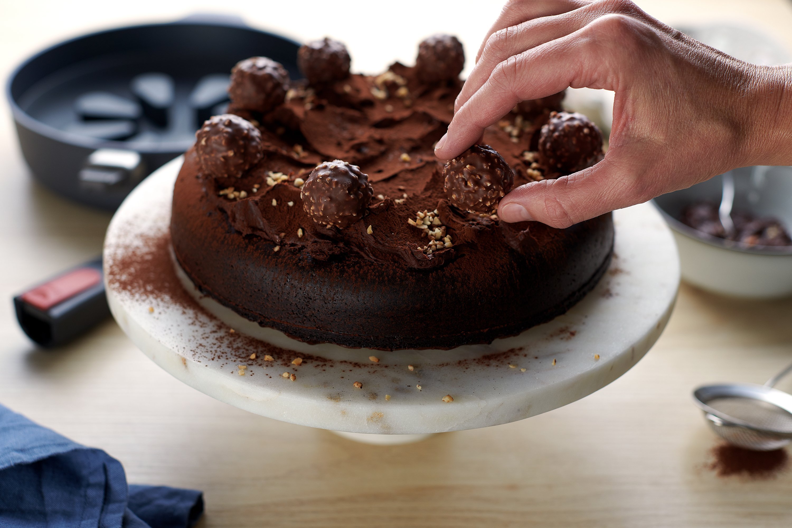 Auf einer Kuchenplatte steht ein frisch gebackener Schokoladen-Haselnuss-Kuchen, der mit Schokoladeganache bestrichen und mit Kakaopulver und Ferrero Rocher dekoriert wird. Im Hintergrund sieht man eine runde Diamond-Lite-Hochrandpfanne mit abgenommenem S