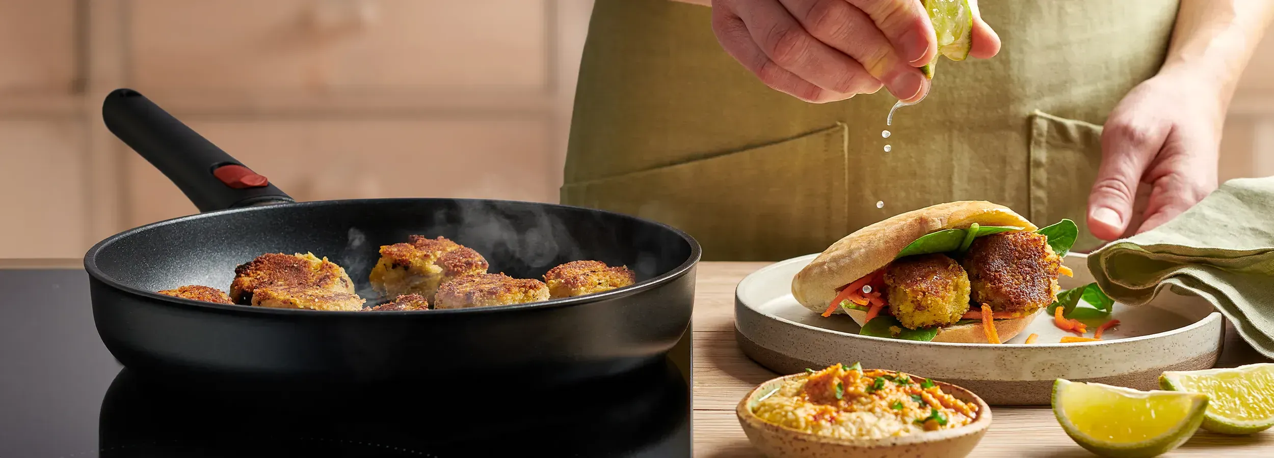 In einer runden, heißen Eco Lite QXR Pfanne werden frische Falafel angebraten. Auf dem Teller daneben liegt ein fertiges Fladenbrot gefüllt mit Falafel und eine Schüssel Hummus steht vor dem Teller.