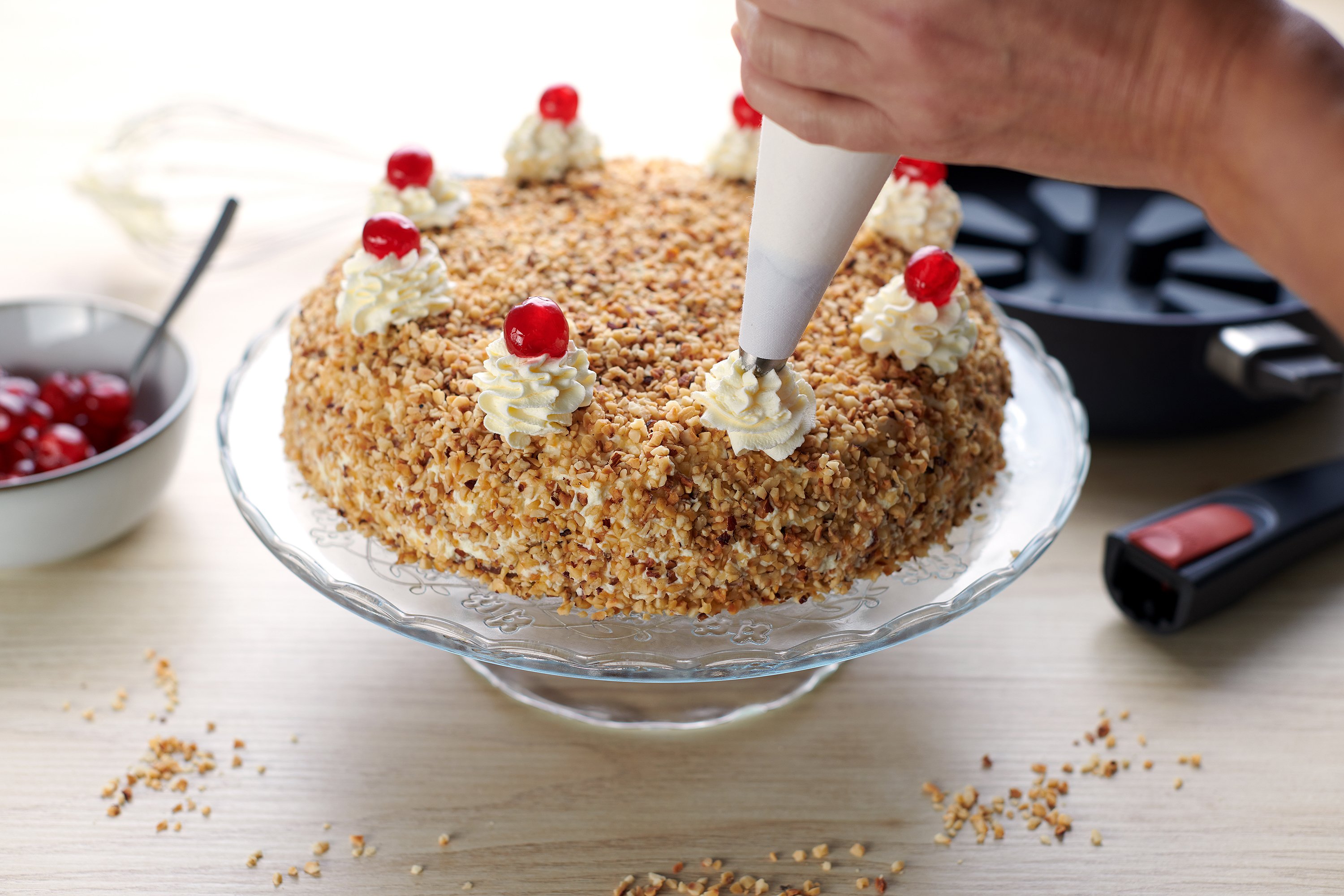 SC24_finished-cake_3000x2000_RGB Auf einer Kuchenplatte steht ein Frankfurter Kranz mit Kirschfüllung. Der Kranz wird mit Sahnehauben und glasierten Kirschen verziert. Im Hintergrund sieht man eine runde Diamond Lite Pfanne mit Silikon-Backeinsatz und abgenommenem Stiel zu sehen.