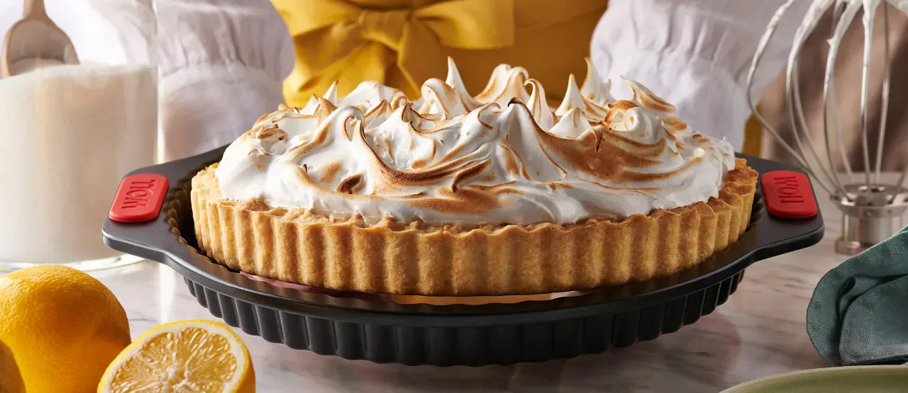 Auf einem Tisch steht eine Quiche- und Tarteform mit Hebeboden aus der Serie Let's Bake! und enthält eine frisch gebackene Zitronen-Baiser-Torte. Daneben liegen aufgeschnittene Zitronen.
