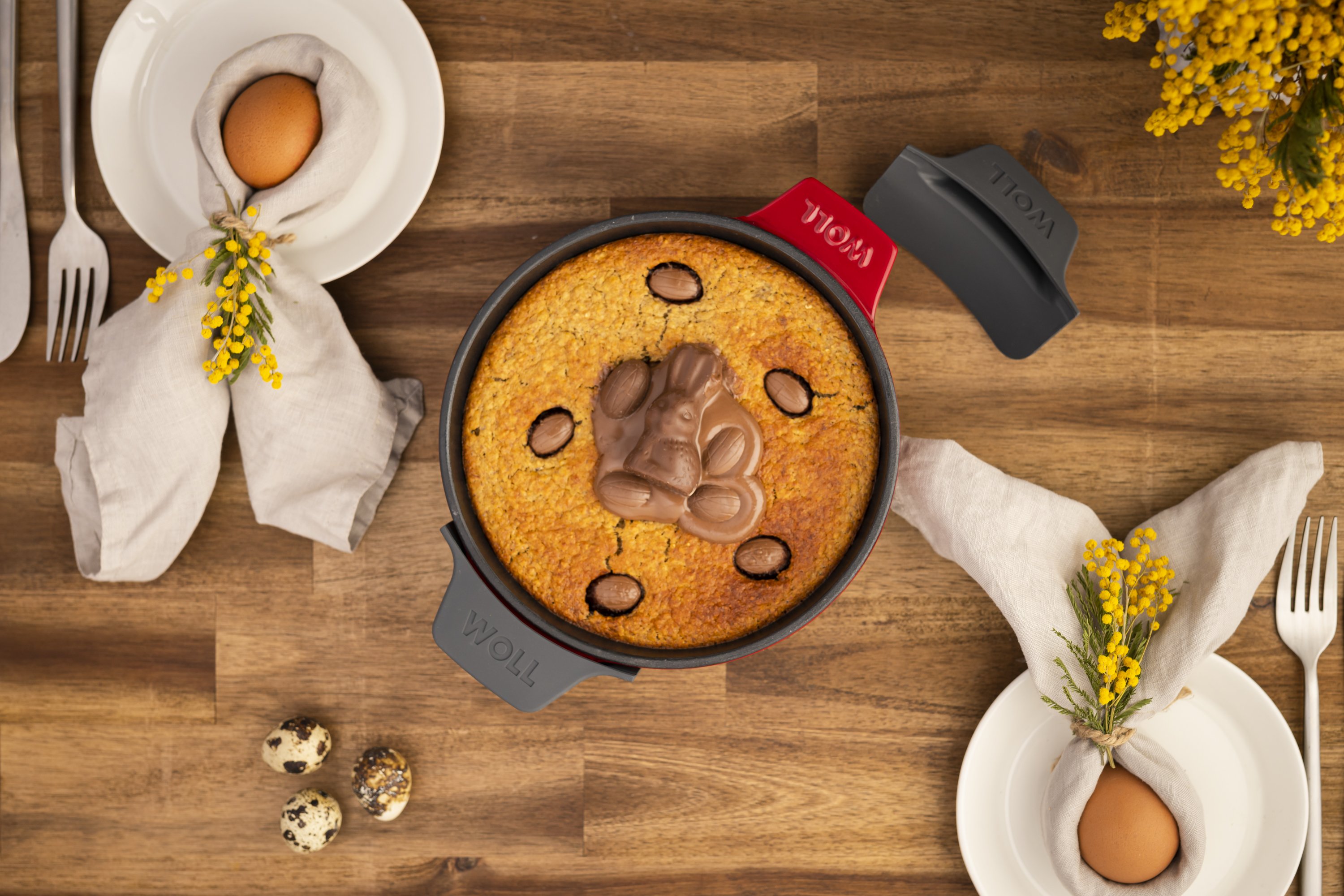 In einer runden Servierpfanne der Serie Iron befindet sich ein frisch gebackener Kuchen mit Schokoladen. Zur Sicherheit wurden Silikongriffe um die Griffe gelegt. Ein Griff liegt daneben, damit die Farbe erkennbar ist. Daneben liegen gekochte Eier auf Tel
