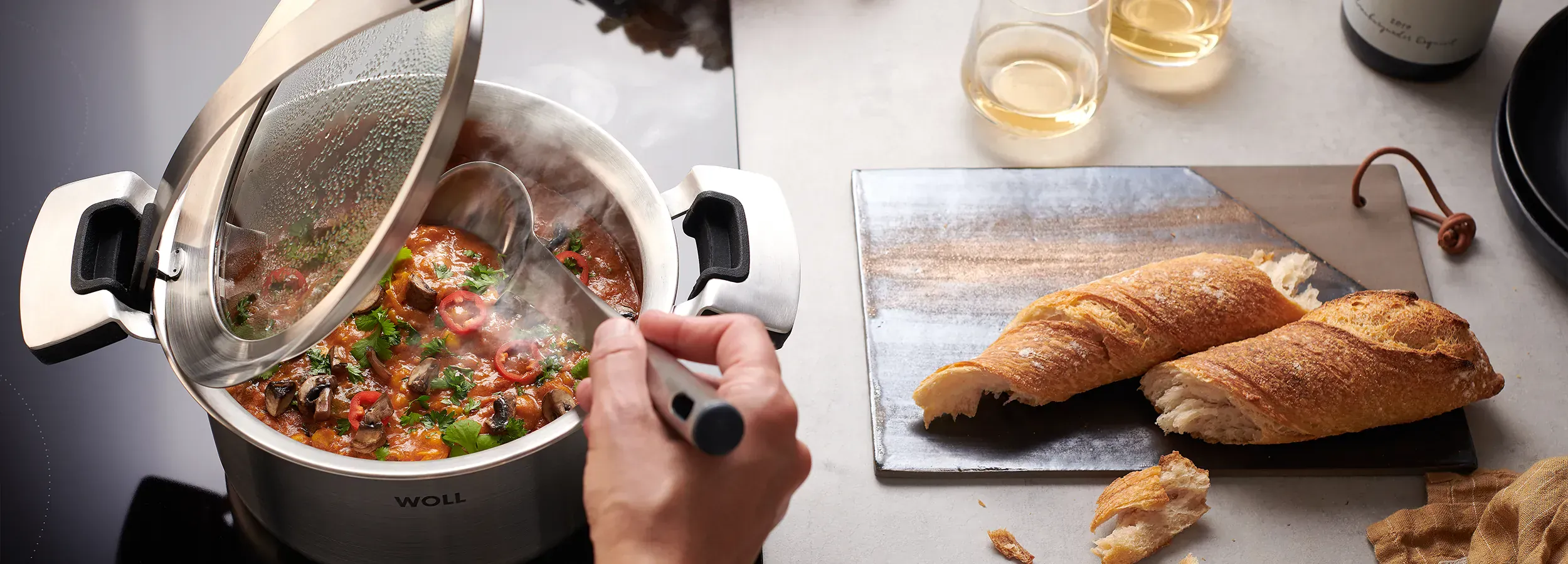 In einem runden Kochtopf der Serie „Concept Pro” wird ein dampfendes Gericht zubereitet. Der Glasdeckel wurde in den Griffeinraster gestellt, sodass er offen bleibt. So kann das Gericht mit dem Kochlöffel umgerührt werden. Rechts daneben liegt ein Schneid