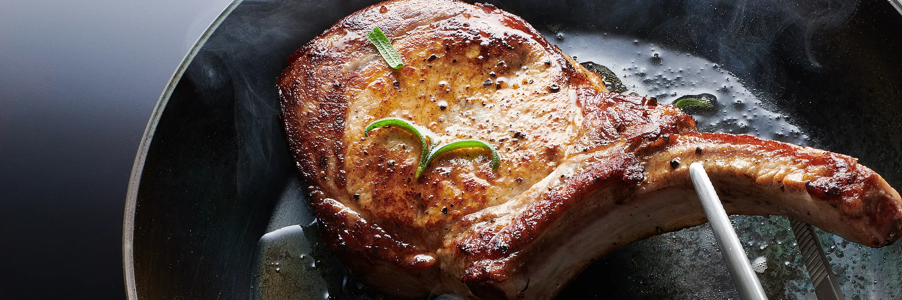 In einer Carbon-Steel-Pfanne mit eingebrannter Antihaftbeschichtung wird ein Stück Fleisch angebraten. Dieses wird mit einer Zange gewendet.