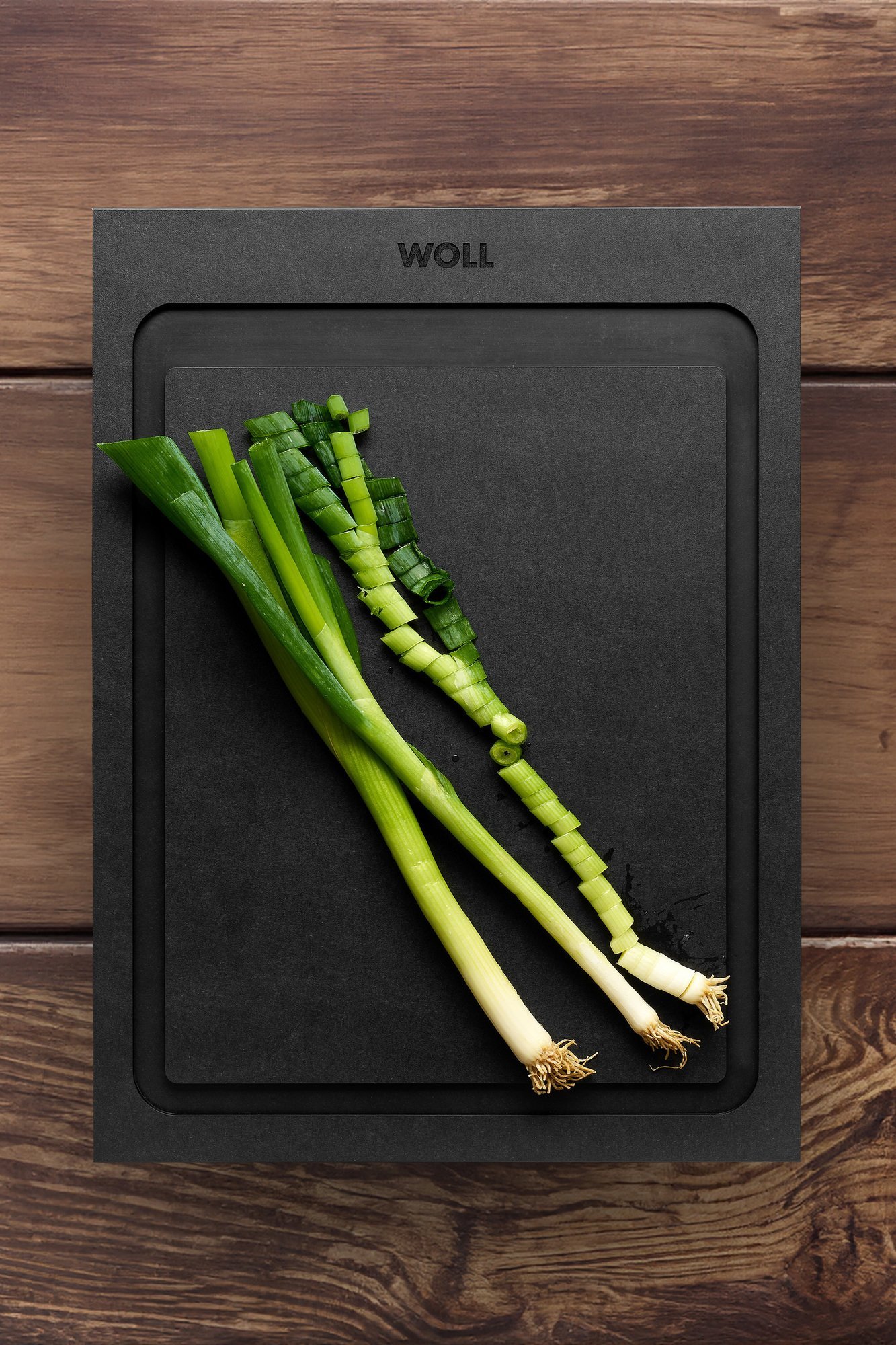 Das Schneidebrett mit Saftrinne der Serie Cut & Serve liegt auf einem Holzbrett. Darauf liegen ganze und geschnittene Frühlingszwiebeln.