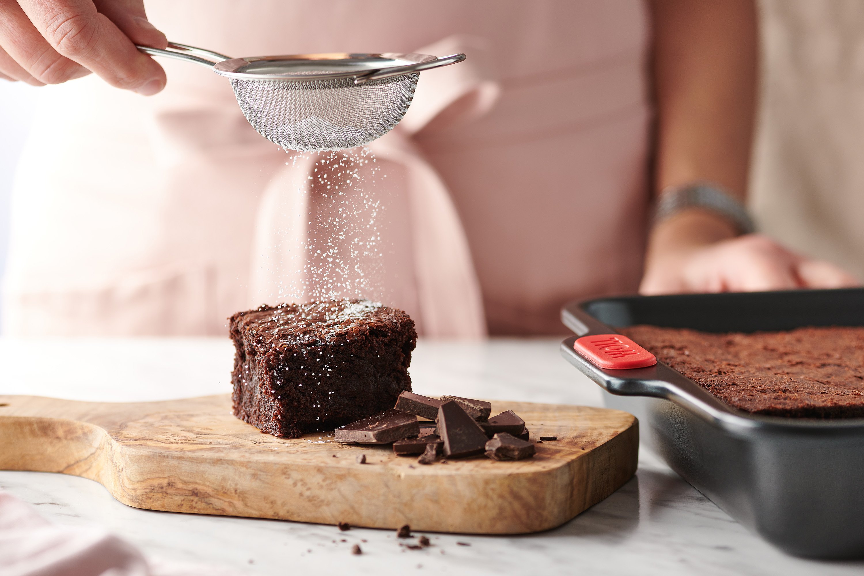 In einer rechteckigen Let's bake! Backform wurden Brownies gebacken. Ein Stück Brownie liegt auf einem Holzbrett und wird mit Puderzucker bestäubt. 