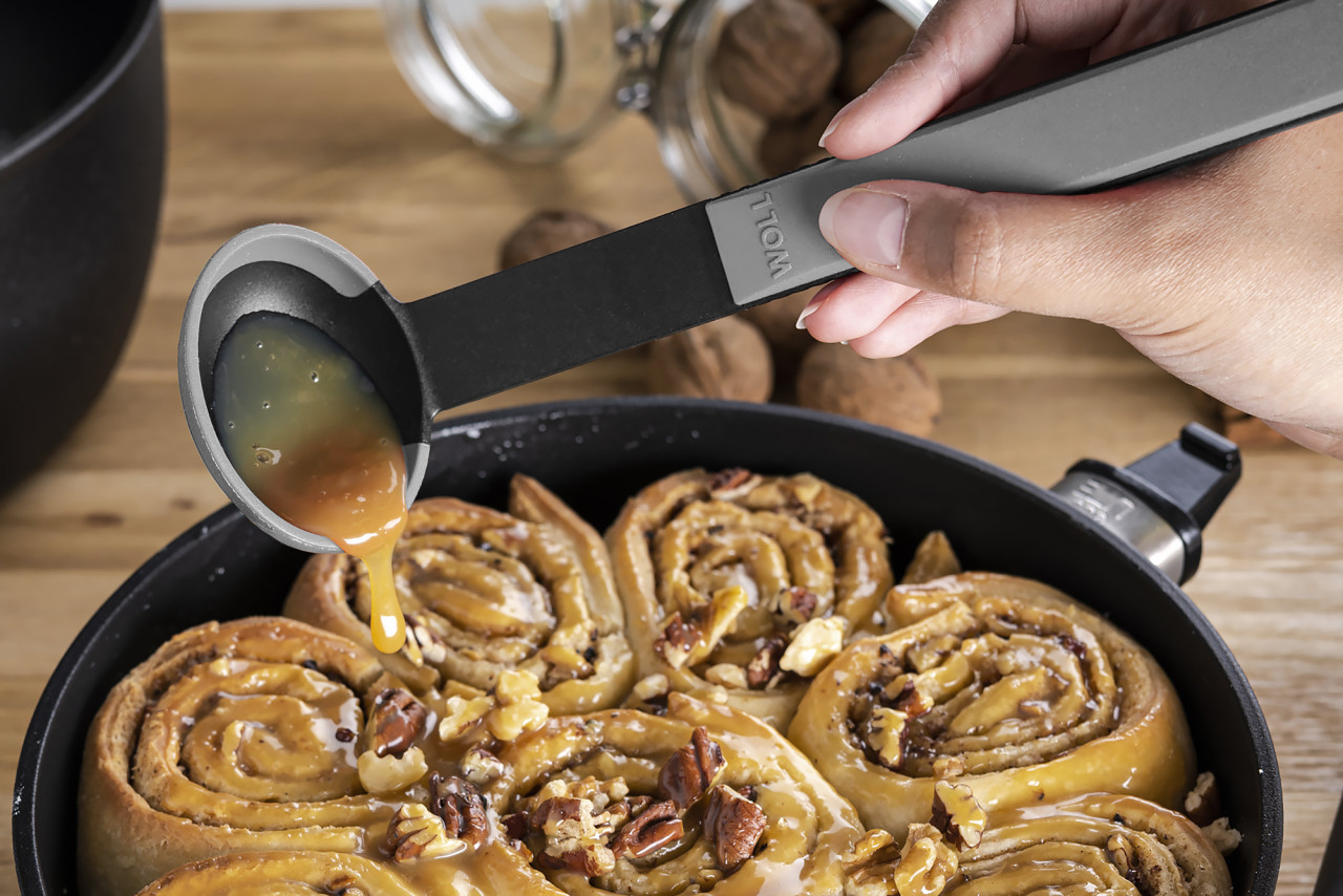 In einer runden Eco-Lite-QXR-Pfanne befinden sich  Zimtschnecken, diese werden mit einem grauen Cock it Saucenlöffel mit Karamellsoße übergossen. 