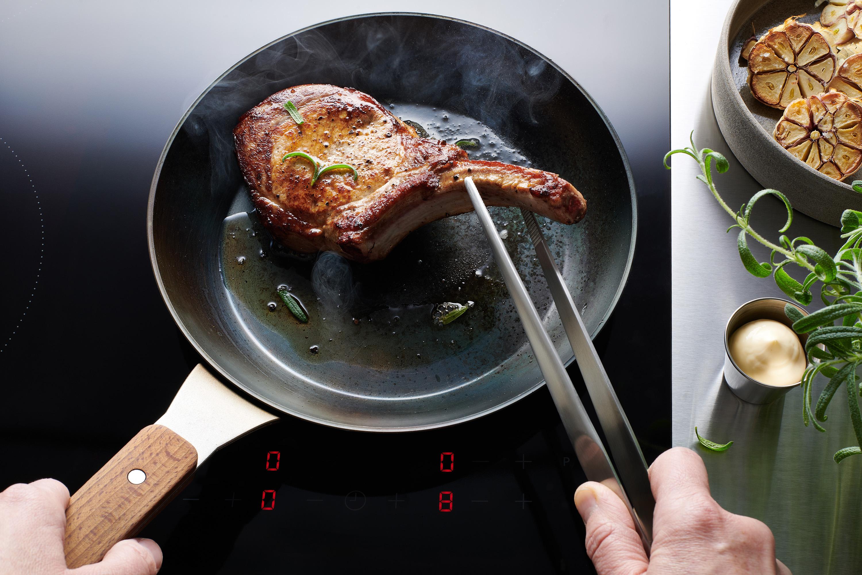 Carbon Steel Flachpfanne In einer runden Carbon Steel Pfanne befinden sich angebratene Steaks. Ein Steak wird auf einem Teller platziert.