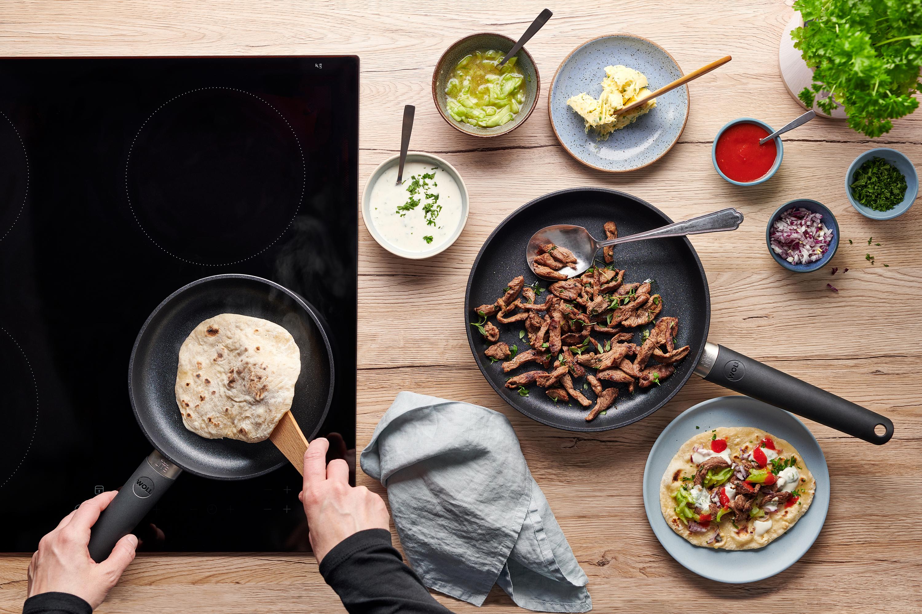 Eco Lite QXR Flachpfanne In der runden Eco-Lite-QXR-Pfanne werden Fladenbrote gebacken. Daneben liegt in einer Pfanne angebratenes Lamm und auf einem Teller ist Döner zubereitet.