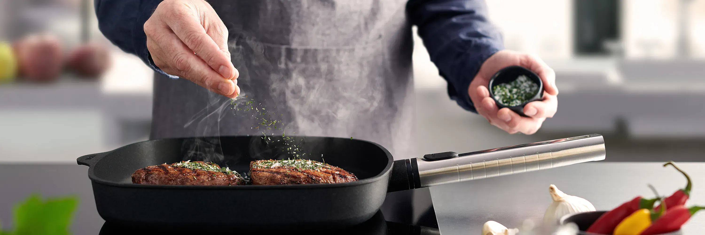 Auf dem Herd steht die rechteckige, heiße Logic-Grillpfanne mit abnehmbarem Edelstahlstiel, in der saftig angebratenes Fleisch dampft. Es ist mit Kräutersalz bestreut. Daneben liegen frische Knoblauchzehen auf der Arbeitsplatte und in einem Behälter stehe