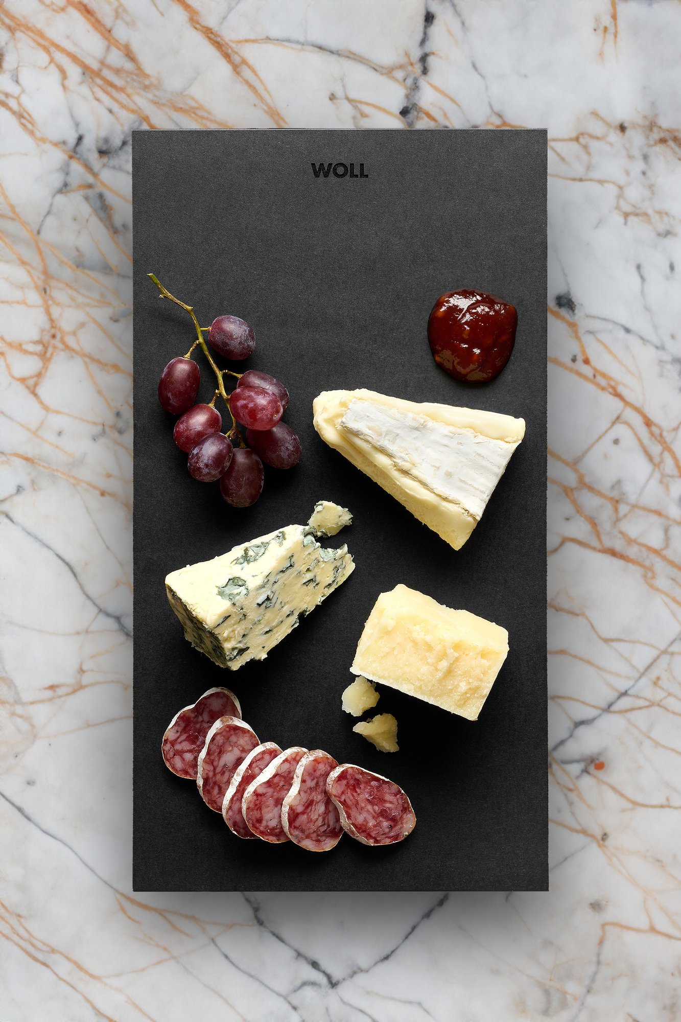 Auf dem Cut & Serve Schneidebrett liegen verschiedene Käsesorte sowie Salamischeiben, Trauben und Marmelade.