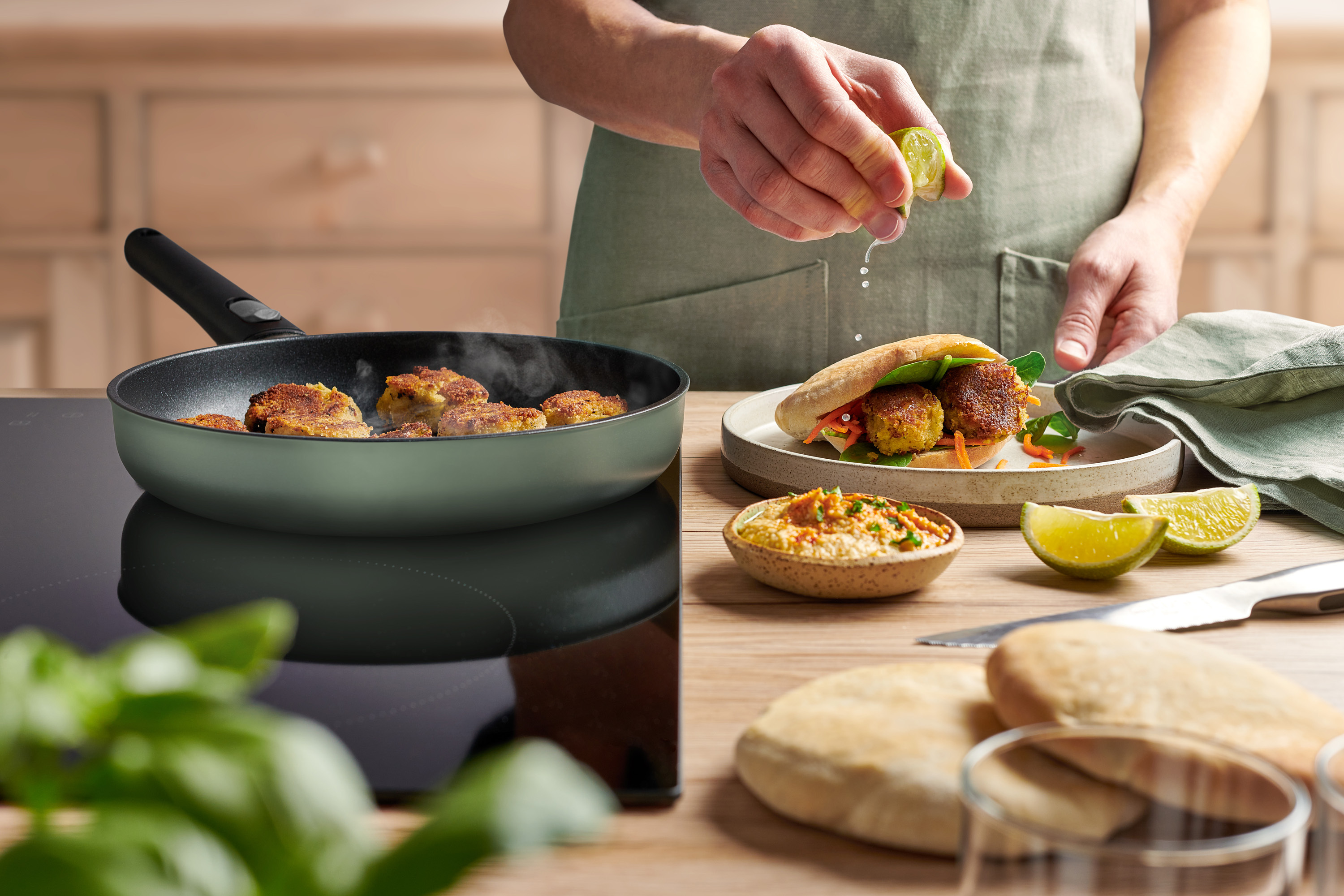 In einer runden, jadegrüne Ecolour Pfanne werden Falafel angebraten. Ein gefülltes Fladenbrot liegt auf einem Teller. 
