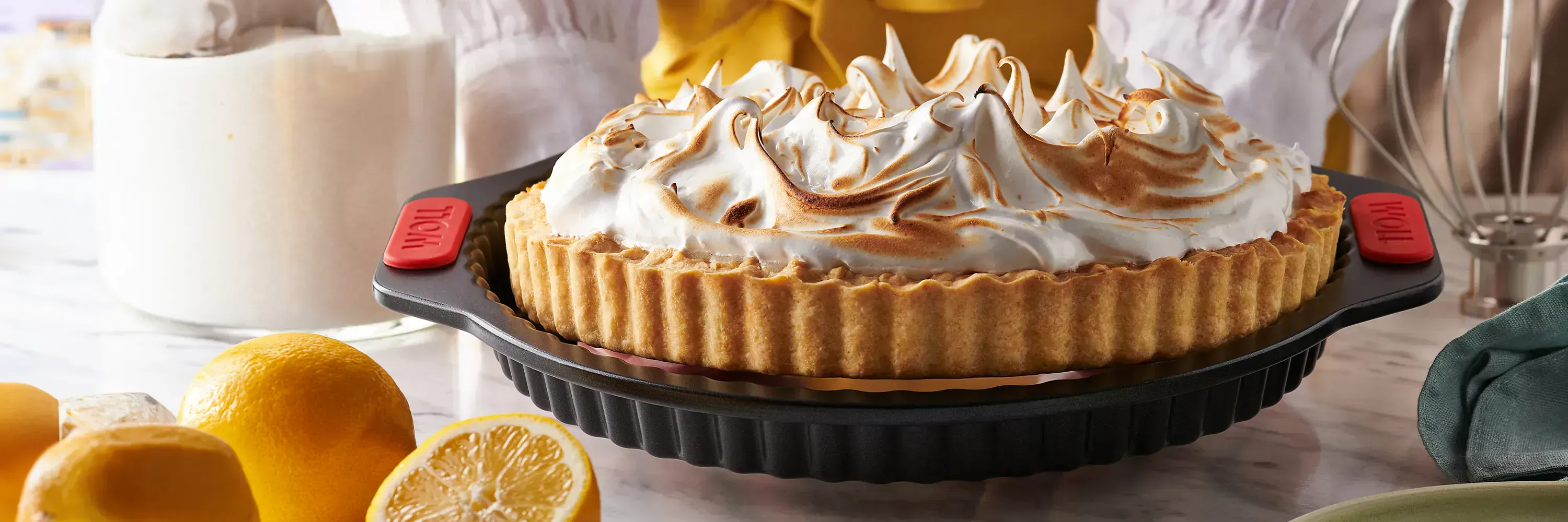 In einer runden Quiche- und Tarteform der Serie „Let's Bake!” befindet sich eine frisch gebackene Zitronen-Baiser-Tarte. Drumherum stehen noch Zutaten wie Zucker und Zitronen sowie ein Rührbesen.