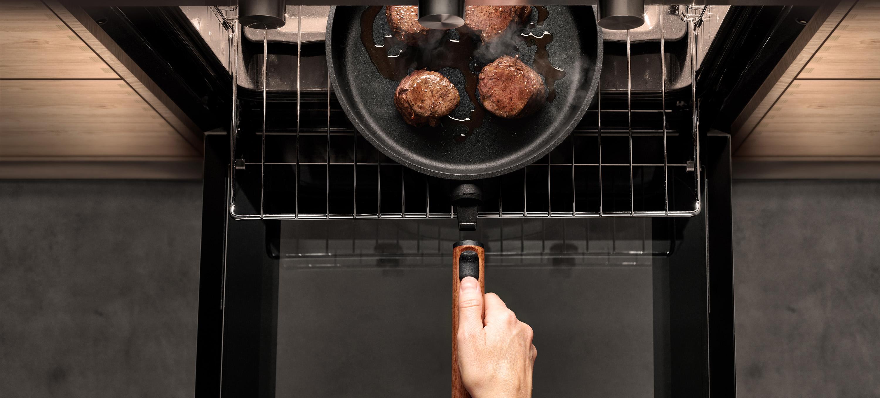 Eco Logic QXR Gusspfanne Auf dem Backofenrost steht eine runde Eco Logic QXR Pfanne mit angebratenem Fleisch. Um die Pfanne zu entnehmen, wird der Stiel eingerastet.