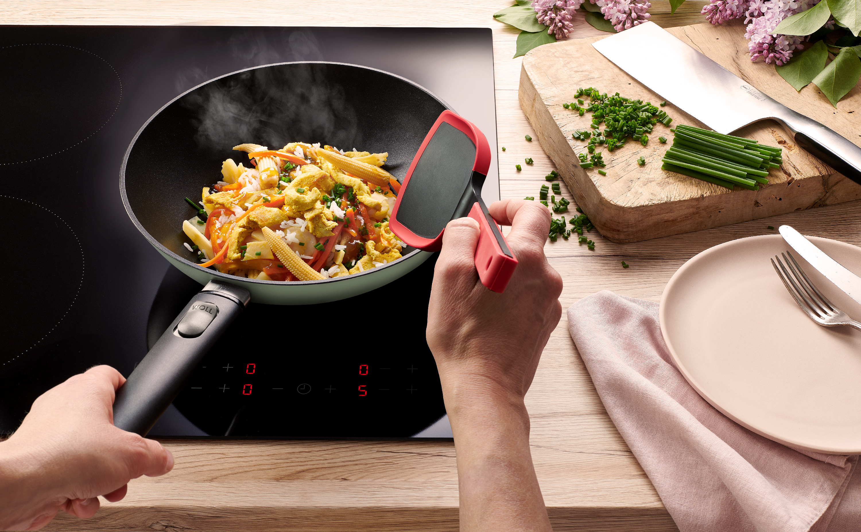 Eine runde, jadegrüne Ecolour Wokpfanne steht auf dem Herd, mit einem Cock it wird ein dampfendes Gericht gewendet. Das Edge Chinesisches Kochmesser liegt daneben.