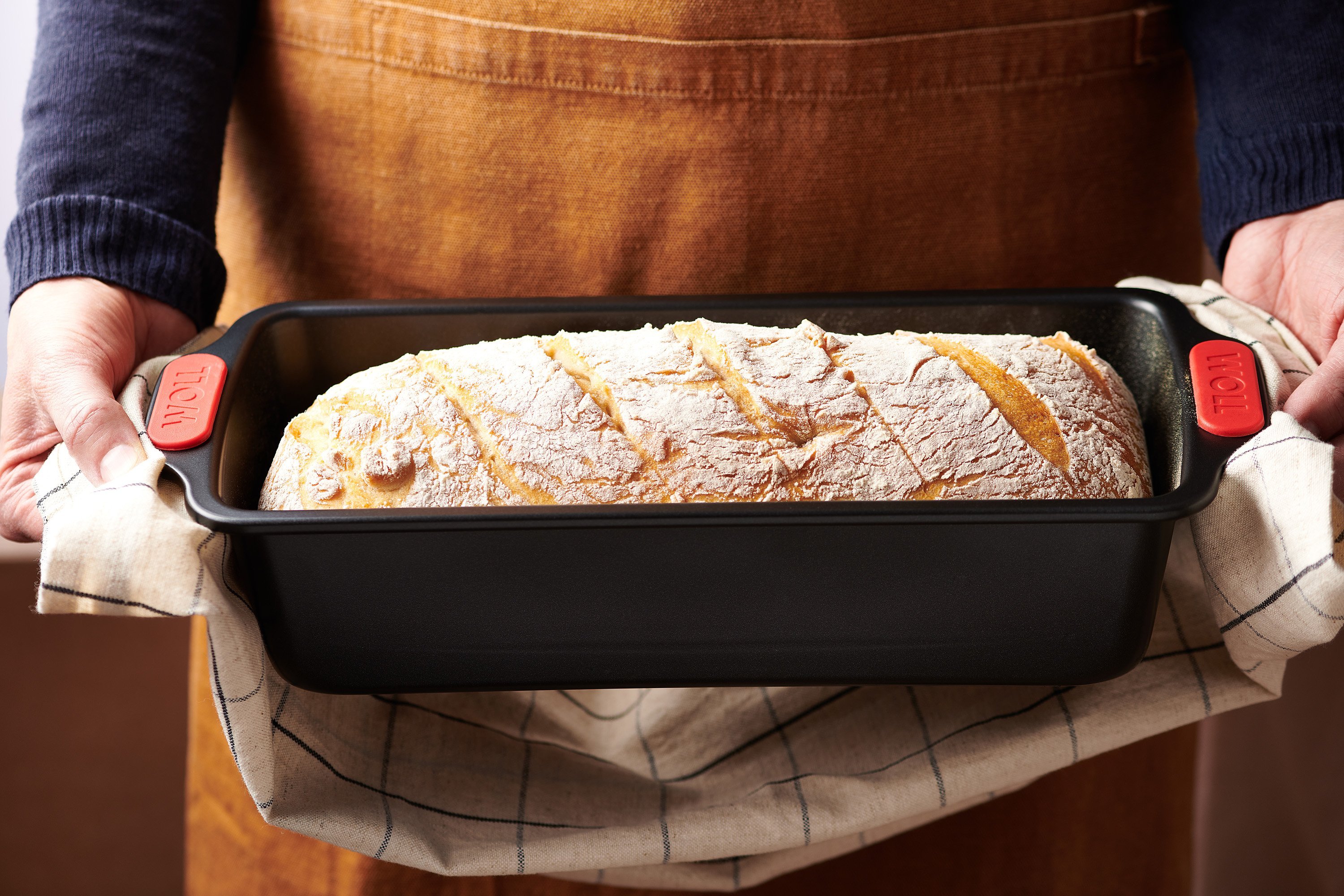 Eine heiße rechteckigen Kastenform der Serie Let's Bake! mit frischem Sauerteigbrot aus dem Backofen wird mit beiden Händen an den Griffen mit einem Küchentuch gehalten.