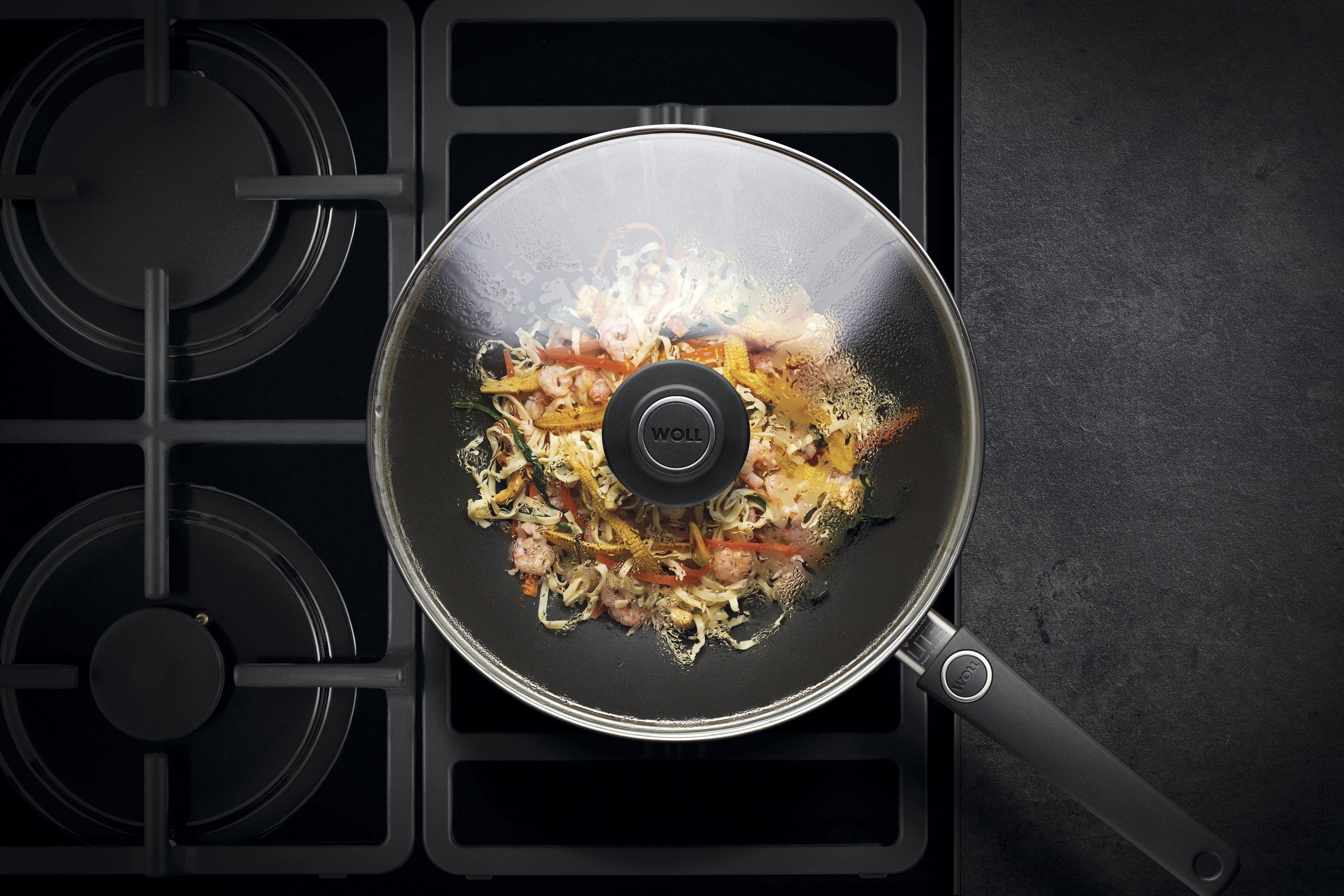 Auf dem Herd steht eine runde, heiße Wok- und Rührpfanne aus der Serie Diamond Lite mit festem Stiel. In dieser befindet sich ein  frisch zubereitetes Gericht. Am Glasdeckel hat sich Kondenswasser gebildet.