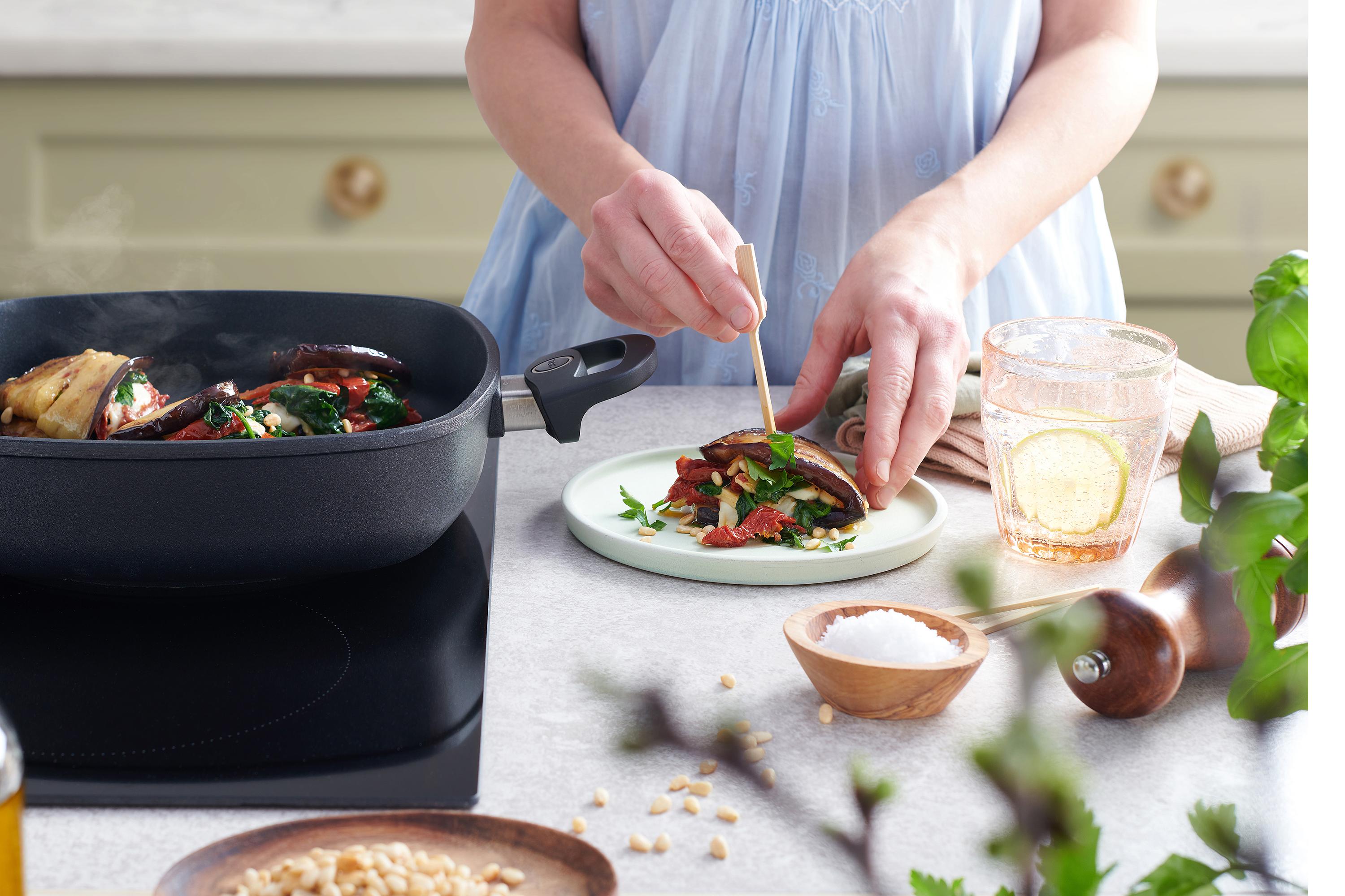 In einer viereckigen Titan Best Pfanne wird ein Gericht angebraten und auf einem Teller daneben angerichtet. 