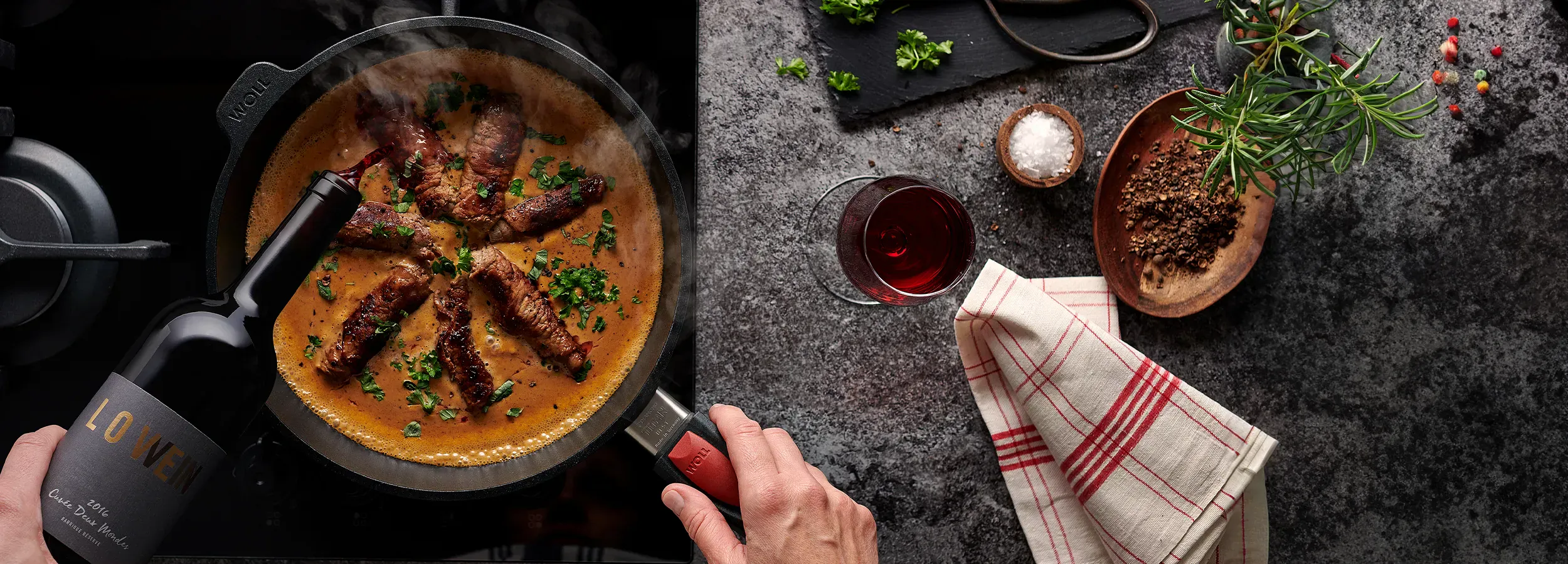 In einer runden, heißen Titan-Best-Pfanne köcheln Rinderrouladen in einer Rotweinsauce. Zur Verfeinerung wird Rotwein hinzugefügt. Auf der Arbeitsplatte befinden sich ein Küchentuch, ein Rotweinglas, eine Schüssel mit Gewürzen und auf einem Brett liegen K