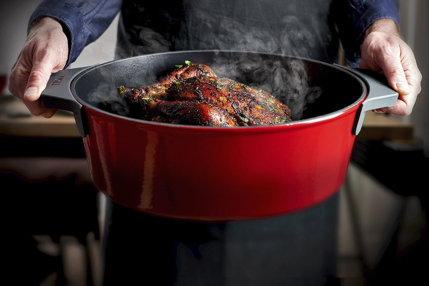 3426CI_V2_Roaster_RGB Der rote Iron Bräuter, in dem sich dampfendes Geflügel befindet, wird gehalten. Zum Schutz vor Hitze werden die Silikongriffe verwendet.