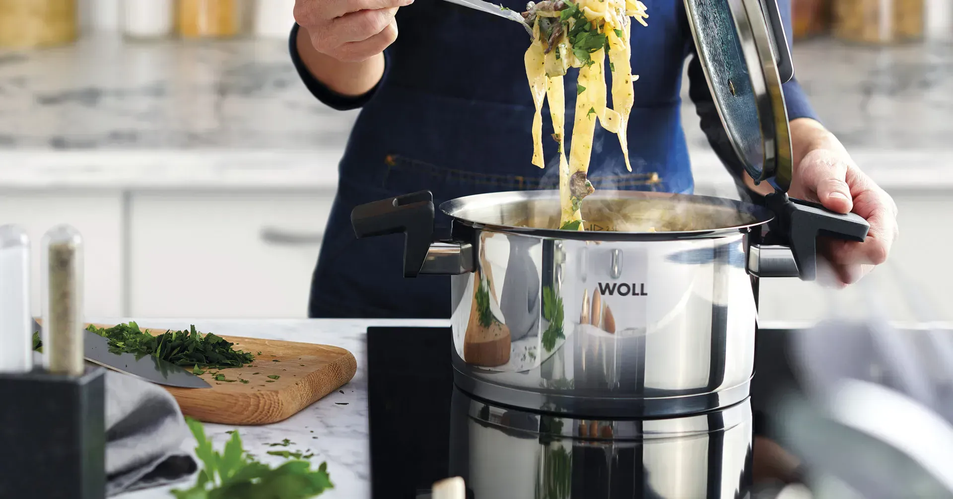 Im runden Edelstahltopf der Serie „Concept” befinden sich frisch zubereitete Bandnudeln. Der Deckel wurde abgenommen und in die dafür vorgesehene Halterung am Griff gesteckt. Daneben liegt ein Schneidebrett mit frischen Kräutern und einem Messer darauf.