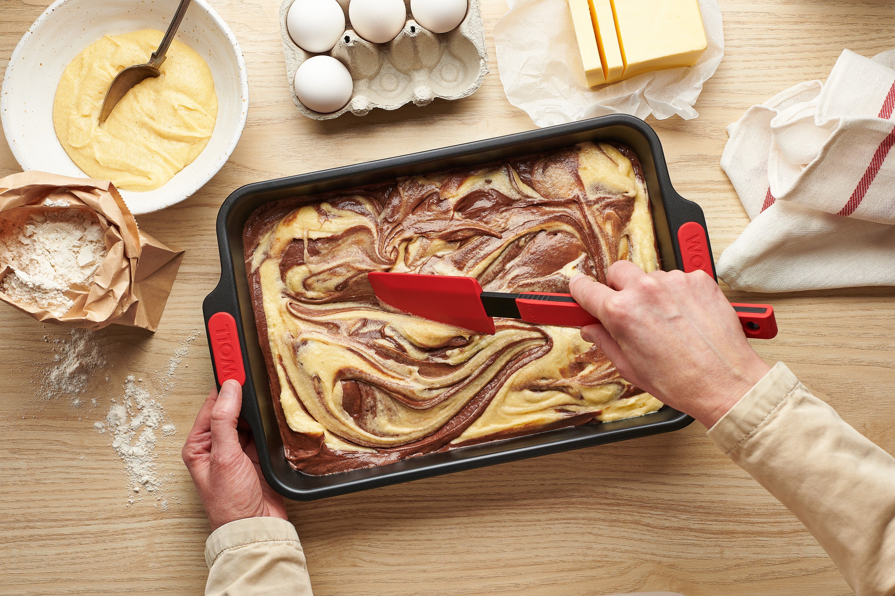 In der Backform der Serie Let’s Bake! befindet sich roher Marmorkuchenteig, der mit einem Teigschaber der Serie Cook i” verteilt wird. Daneben sind eine offene Packung Mehl, ein Küchentuch sowie eine Schale mit Teig zu sehen.